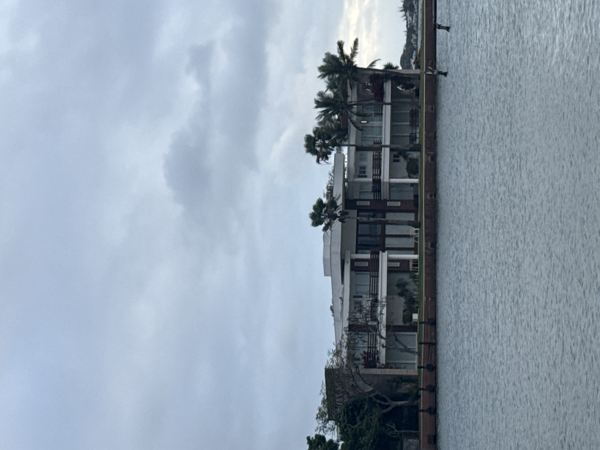 This image shows a modern, multi-story house located on the waterfront in Cabo Frio, Brazil. The house is characterized by its white and dark brown exterior, multiple balconies with railings, and large glass windows. Several palm trees are visible in front of and around the house, suggesting a tropical or subtropical climate. The foreground features a body of water, likely the sea or a large bay, with some wooden posts or pilings visible in the water near the shore. The sky is overcast with light clouds, indicating a possibly gloomy or hazy day. The overall impression is one of a luxurious waterfront property in a coastal city.