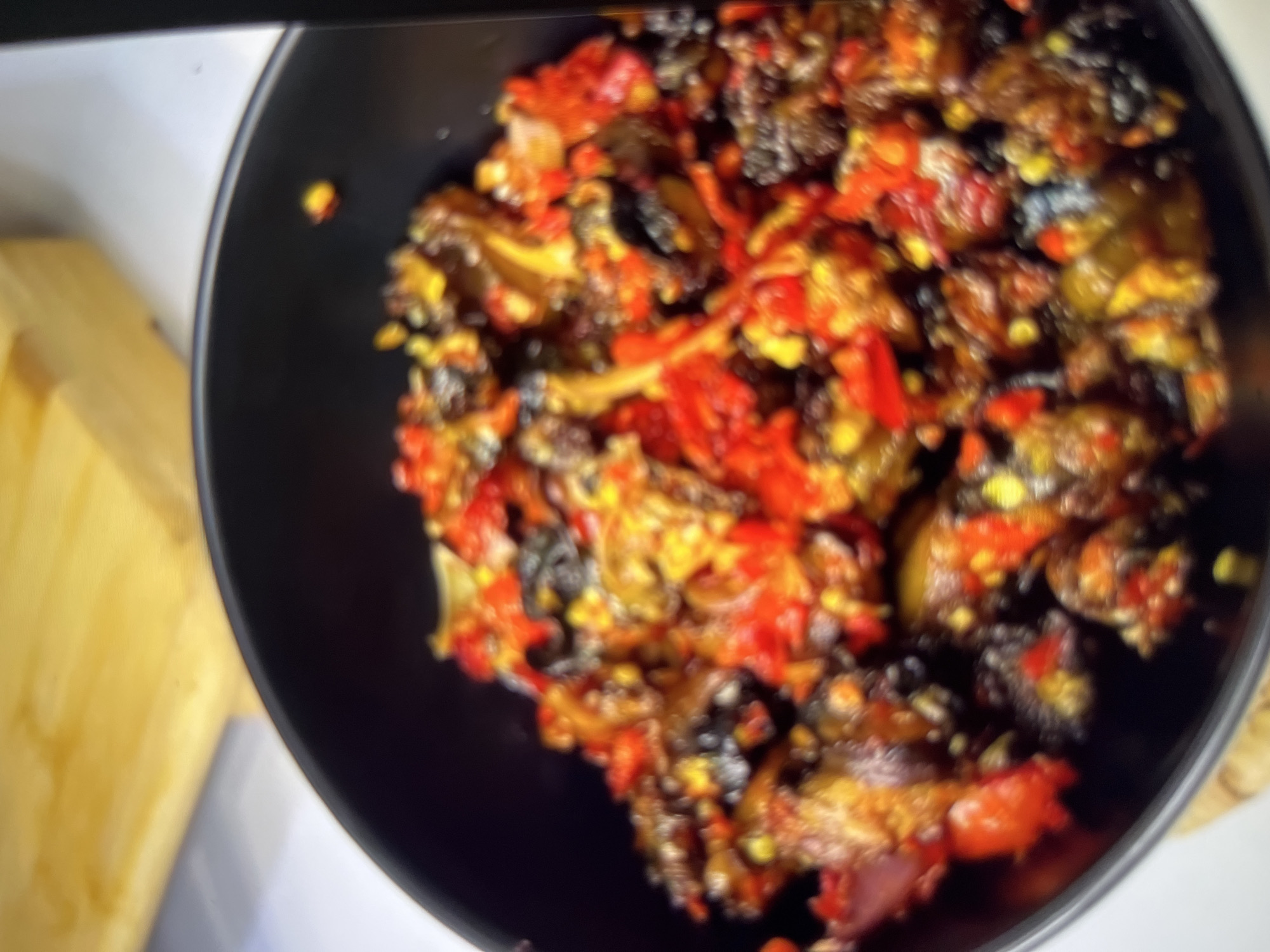 The image shows a close-up, slightly blurry view of a black bowl filled with what appears to be a spicy stew or dish. The dish is composed of dark, chunky pieces mixed with vibrant red chili peppers and yellow flecks, possibly corn or other vegetables. The lighting suggests an indoor setting, with a hint of a wooden surface in the upper left corner, possibly a countertop or cutting board. No specific location in Abuja, Nigeria, can be identified from the visual information provided.