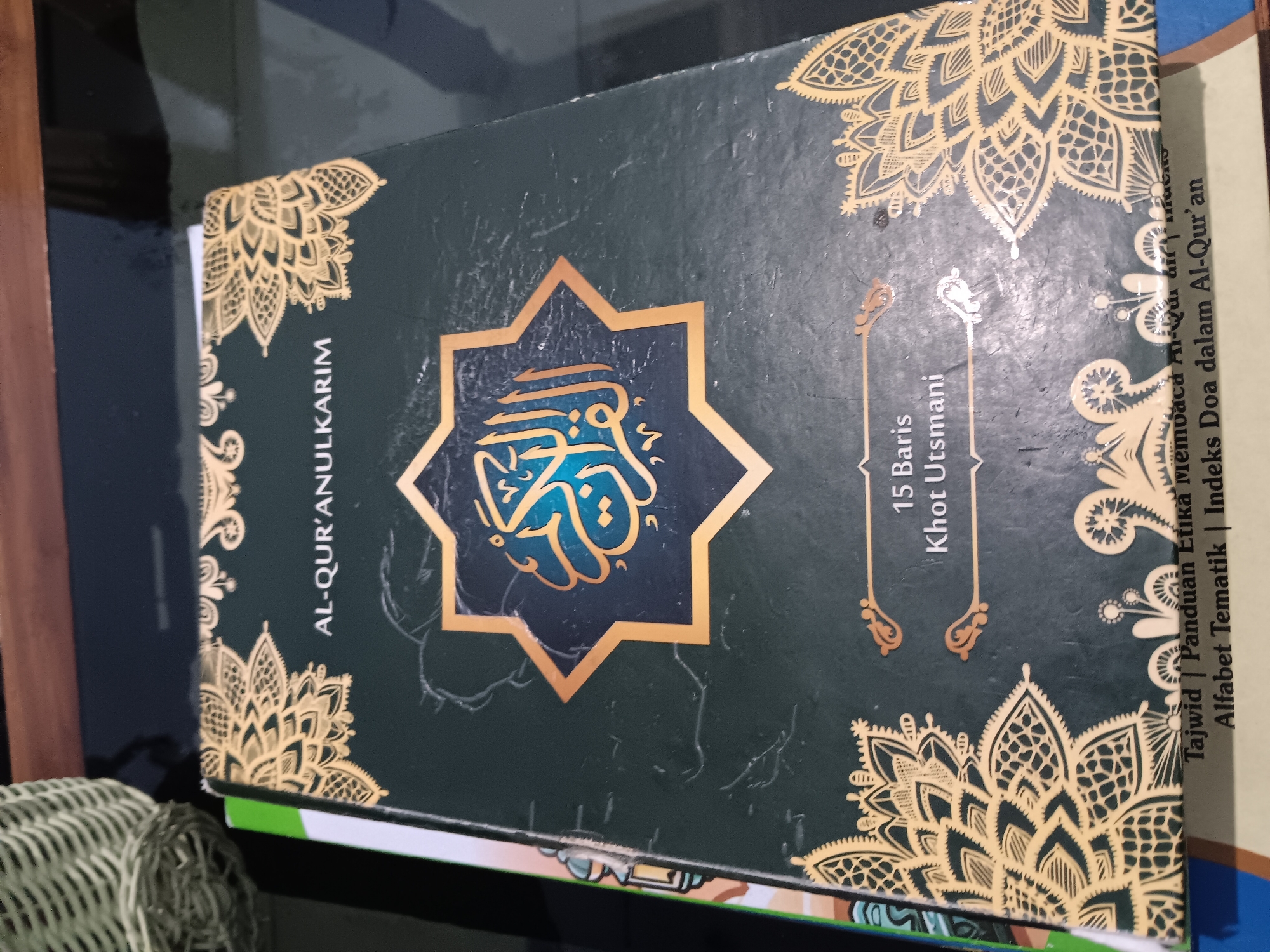 The image shows the cover of a dark green book, presumably a copy of the Quran, with ornate gold patterns. The title "AL-QUR'ANULKARIM" is prominently displayed in gold lettering above a central, star-shaped motif containing Arabic calligraphy. Below this, in smaller gold text, are the words "15 Baris" and "Khot Utsmani". At the bottom of the cover, more gold lettering indicates content such as "Tajwid | Panduan Etika Membaca Al-Qur'an" and "Alfabet Tematik | Indeks Doa dalam Al-Qur'an". The book is lying flat on a reflective, dark surface, possibly a table. To the left of the book, part of another book or item with a green and white cover is visible. In the upper left corner, a portion of a woven basket is present. The image is taken from a slightly elevated angle, focusing on the book's cover. The overall lighting suggests an indoor environment. The specific location in Batursari, Indonesia, is not visually ascertainable from the image.