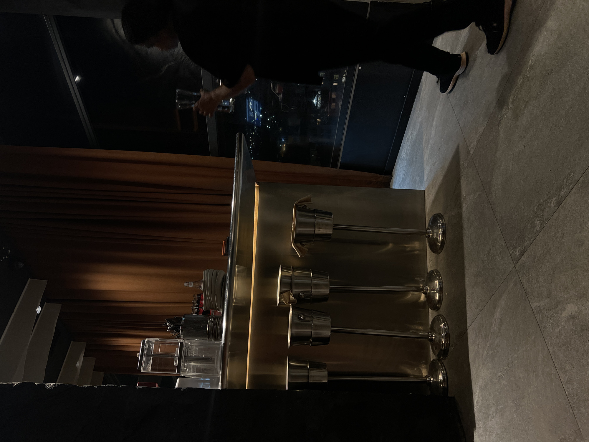 A person in black stands at a bar in Yerevan, Armenia. The bar is silver with multiple bar stools. A window overlooks the city.