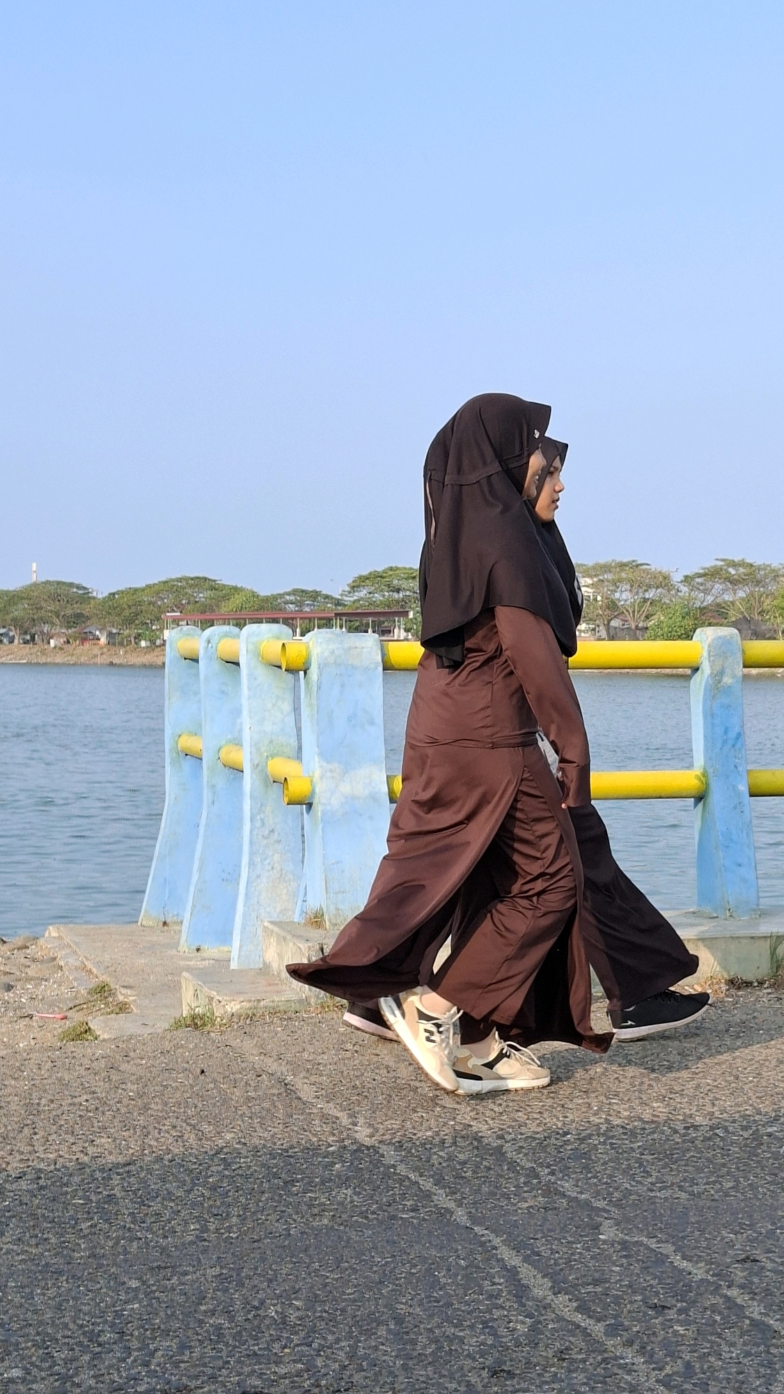 Two women wearing brown and black hijabs and long brown dresses walk along a paved path beside a body of water. A blue and yellow barrier separates the path from the water. Trees and buildings are visible in the background. The scene appears to be at the TPI Lhokseumawe, a fishing port in Lhokseumawe, Indonesia.
