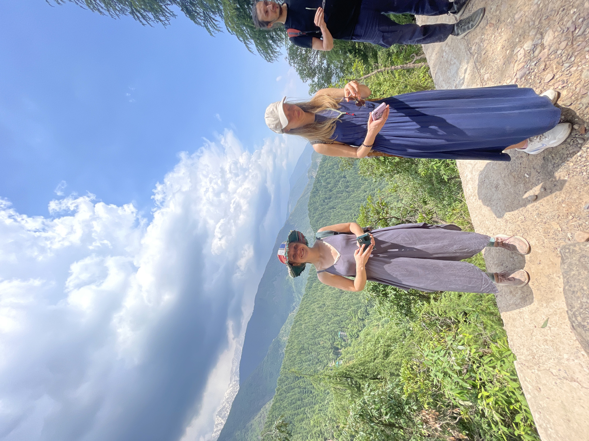 A daytime, landscape-oriented photograph captures a group of three individuals on an elevated, concrete ledge in Dharamkot, India, overlooking a mountainous terrain.
The woman in the foreground wears a patterned hat and a dark jumpsuit, using a mobile phone. A second woman stands behind her, dressed in a long blue dress and a light-colored baseball cap, also holding a phone. Further back, a man in a black shirt and pants stands slightly obscured.
The backdrop consists of lush, green mountain ranges under a partly cloudy sky. Some areas of the mountain display visible signs of snow. The concrete ledge provides a distinct foreground, contrasting with the natural scenery.