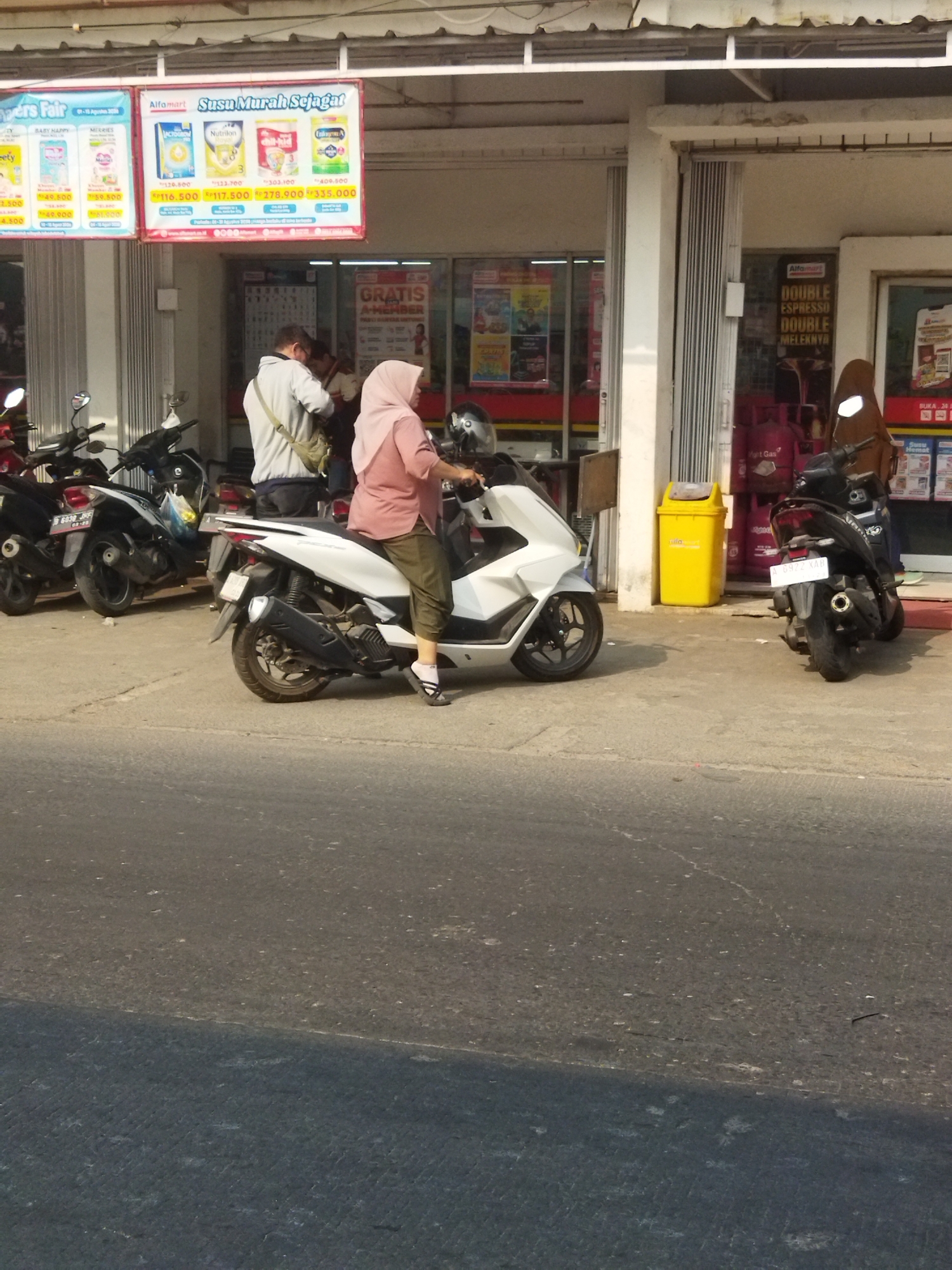 The image depicts the exterior of an Alfamart convenience store in Sepatan, Indonesia, on a sunny day. In front of the store, several motorcycles are parked on a paved area, with a white scooter in the foreground occupied by a woman wearing a pink hijab and olive-green pants. She is looking to her right. Behind her, a man in a grey jacket and a beige backpack stands near the store entrance. The store's facade features promotional banners, including one advertising "Susu Murah Sejagat" (Cheap Milk Everywhere) and another with "GRATIS A-MEMBER" (Free A-Member). Gas cylinders are visible inside the store. The overall environment suggests a typical street scene in an Indonesian town, with commercial activity and everyday life.
