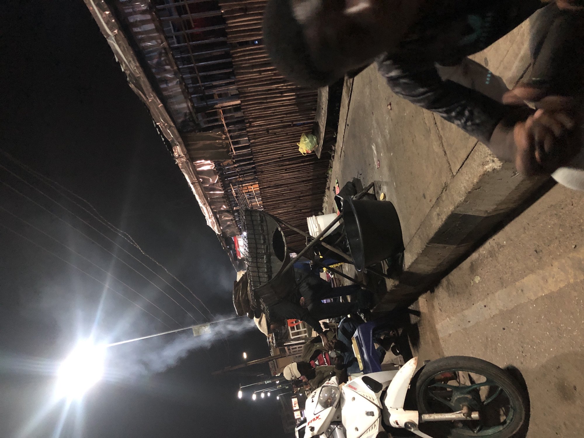 The image depicts a nighttime scene in Jos, Nigeria, likely at a street market or roadside gathering. Several people are visible, some interacting with each other and others focused on their tasks. In the foreground, two motorcycles are parked, one predominantly white with visible branding like "FXR". Smoke or steam drifts across the scene, illuminated by a bright light source, possibly a street lamp or a vehicle's headlight.
The setting appears to be outdoors, with a paved ground surface and a makeshift structure overhead, possibly part of market stalls or temporary roofing, constructed from wood and reflective material. The lighting is dim, characteristic of nighttime, with bright artificial lights creating strong contrasts and shadows. The overall atmosphere is busy and informal.
One person is wearing a helmet. Another individual appears to be handling something on a table or stand. The wooden structure above has a somewhat ramshackle appearance, suggesting a temporary or informal setup. The presence of the motorcycles and the general ambiance suggest a lively roadside scene where people gather, perhaps for commerce or socializing. The time of day is clearly night, and no specific weather conditions are discernible beyond the presence of the smoke or steam. There are no clearly legible text elements on the visible objects or clothing.