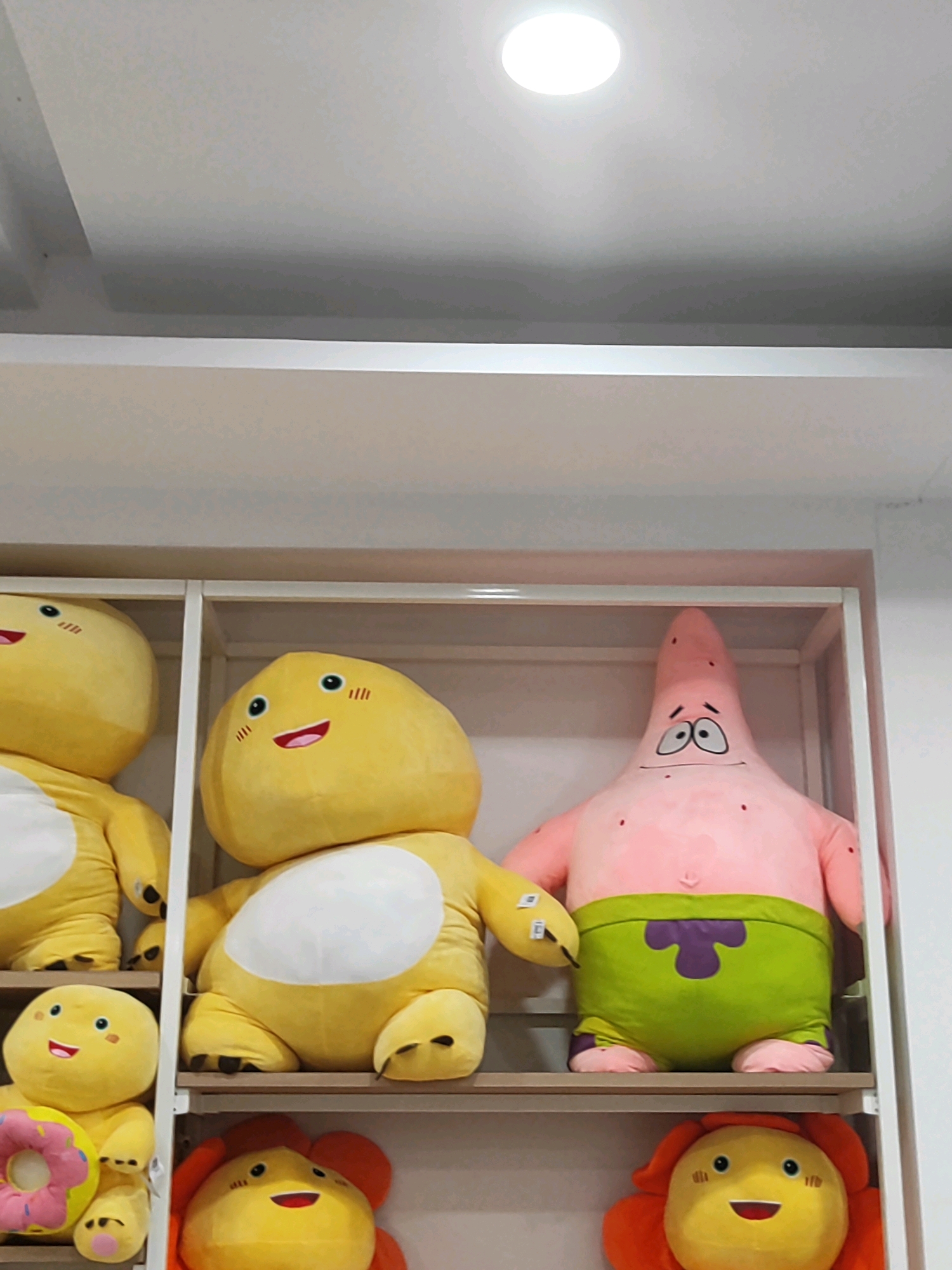 In Sukowiryo, Indonesia, three shelves of stuffed toys are visible. The top shelf displays a large yellow stuffed toy on the left and a pink starfish-shaped stuffed toy, resembling Patrick Star from SpongeBob SquarePants, on the right. The middle shelf holds a yellow stuffed toy with a round head and body on the left, and a large yellow stuffed toy with a similar round head and body on the right. The bottom shelf features a yellow stuffed toy with a flower on the left and another yellow stuffed toy with a flower on the right. The stuffed toys are arranged on white shelves within a white shelving unit. A circular light fixture is visible on the white ceiling above.