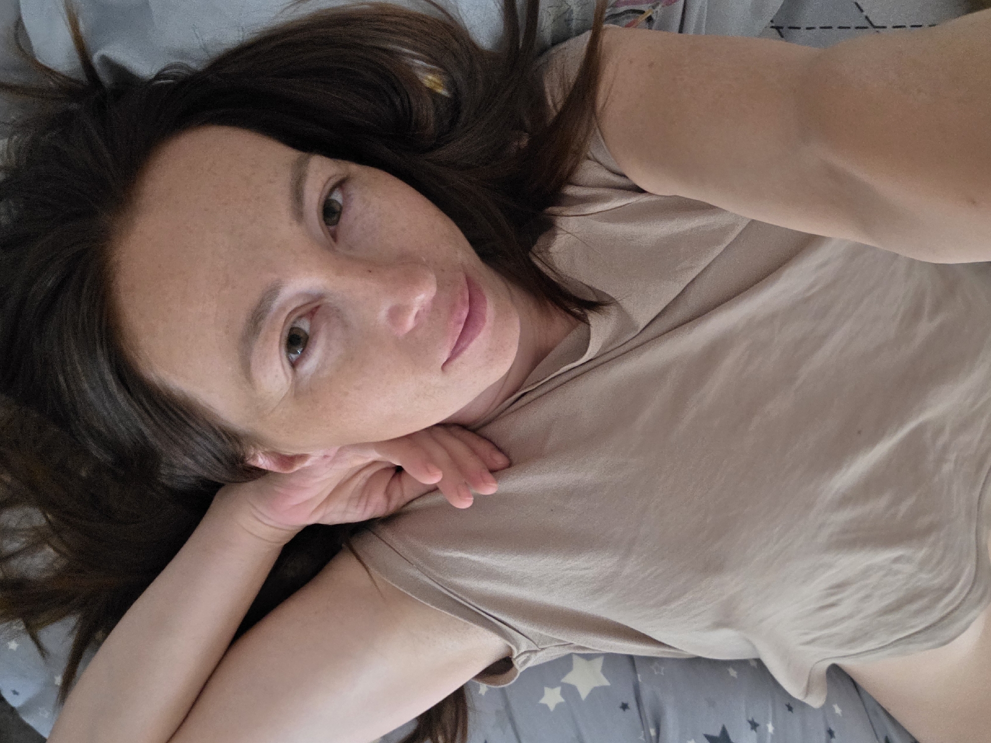 A woman is lying in bed, taking a selfie. She is wearing a beige t-shirt and has long dark hair. Her eyes are green and she is looking at the camera. The bed has a gray sheet with white stars. The image was taken in Chaikovsky, Russia.