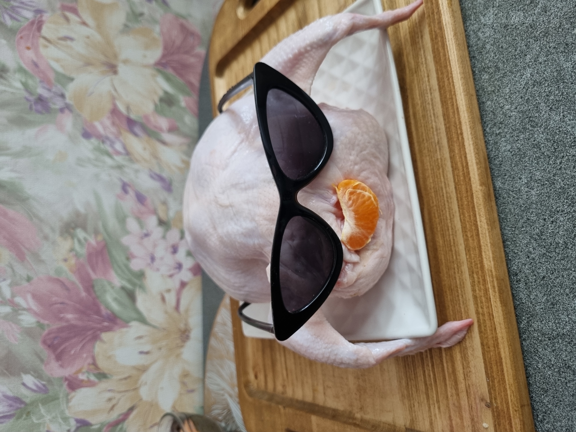 The image displays a raw, whole poultry, likely a chicken, situated on a white rectangular dish. The poultry is humorously accessorized with black cat-eye sunglasses placed over the "eyes" area and a peeled tangerine segment positioned where the mouth would be. The dish rests on a light brown wooden cutting board with parallel grooves. Beneath the cutting board is a grey, textured surface. In the background, a wall is covered with wallpaper featuring a floral pattern of large blooms in shades of pink, yellow, and purple against a lighter background. A portion of a clear glass containing a dark liquid is partially visible on the far left. The scene is located in a kitchen setting in Chaikovsky, Russia.
