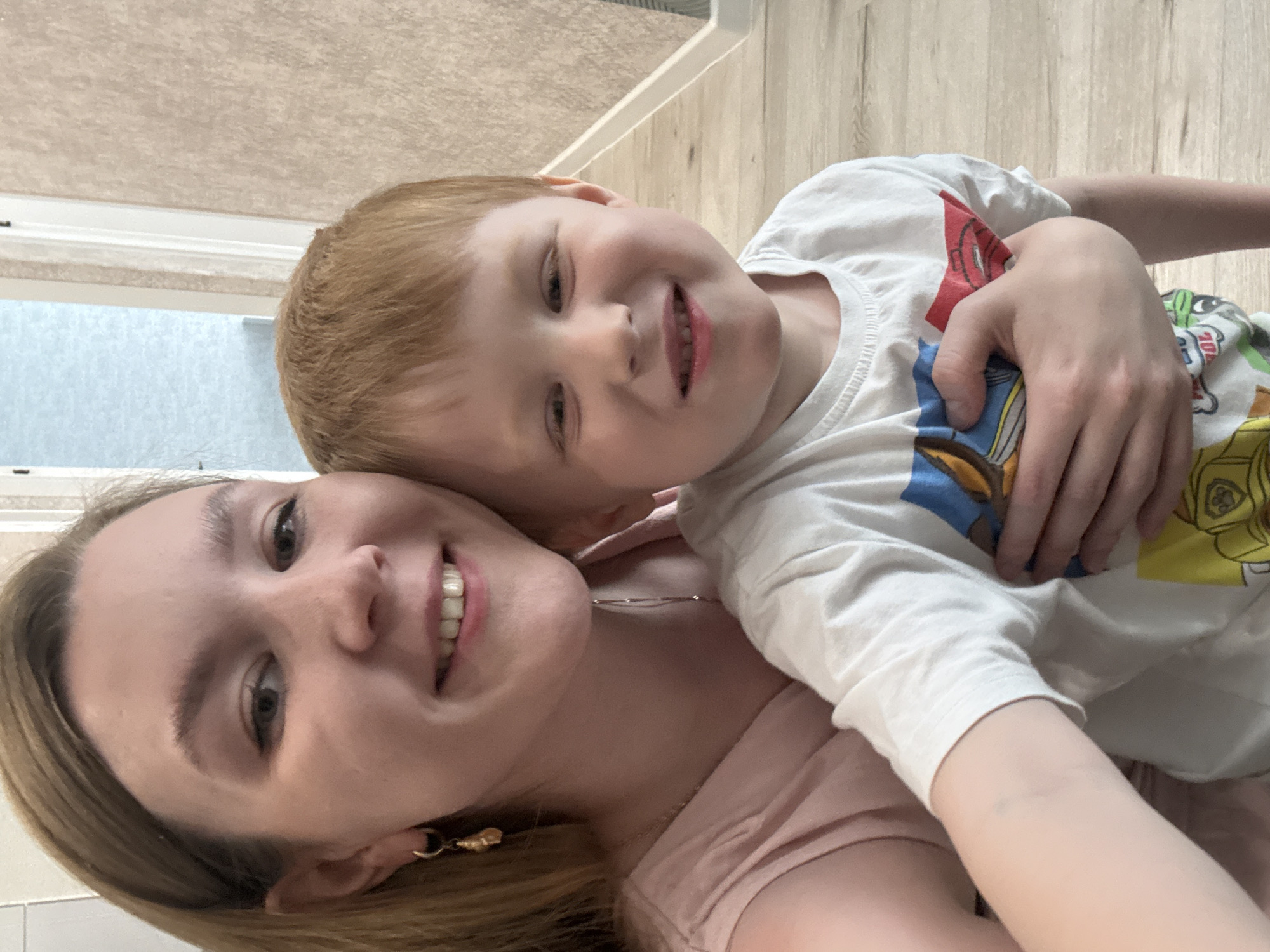 A selfie of a woman and a young boy is oriented at an angle with the woman on the left and the boy on the right. Both are smiling at the camera. The woman has fair skin, blonde hair, and is wearing a pink top and gold earrings. The boy has fair skin, red hair, and is wearing a white long-sleeved shirt with a cartoon pattern. The boy's arm is wrapped around the woman. The background includes a neutral-colored wall on the left and a wooden floor on the right. There is no indication of the location being Izhevsk, Russia.