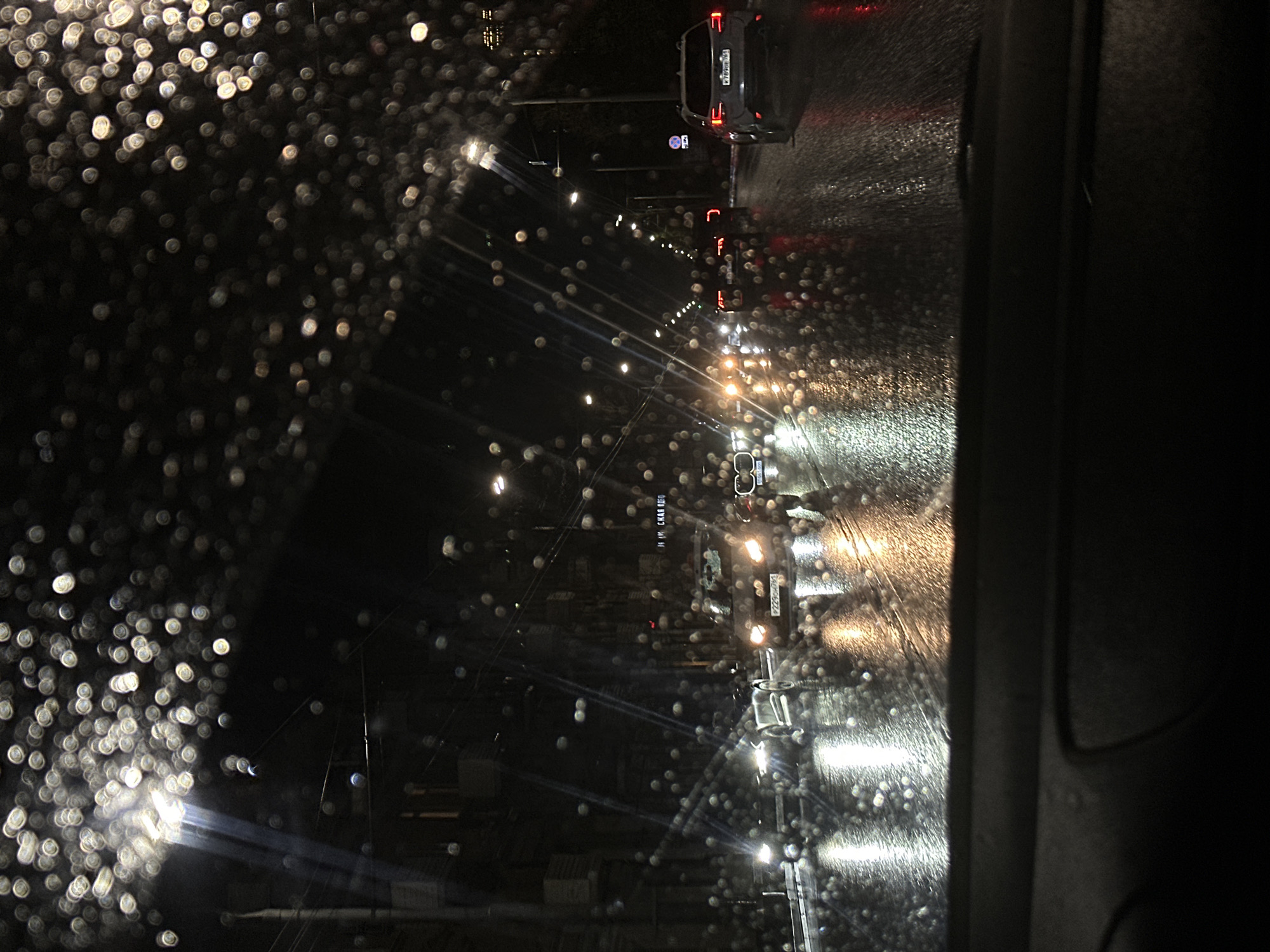 The image shows a rain-streaked window with blurred lights and a car in the background. The perspective is from inside a vehicle, looking out onto a wet city street at night. The license plate on the car behind is readable in the full image and shows a location in Russia, Ovoshchnoy. The raindrops on the window distort the city lights into blurred circles. The street appears to be moderately busy, with several other vehicles visible in the distance.