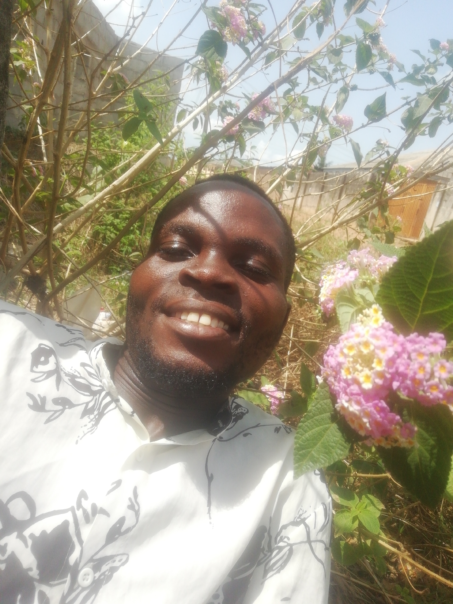 The image shows a smiling man in a white shirt with a black floral pattern. He appears to be in an outdoor setting with flowers and greenery surrounding him. There are tree branches in the background and a building structure is partially visible. The location is in the city of Unknown, Unknown.
