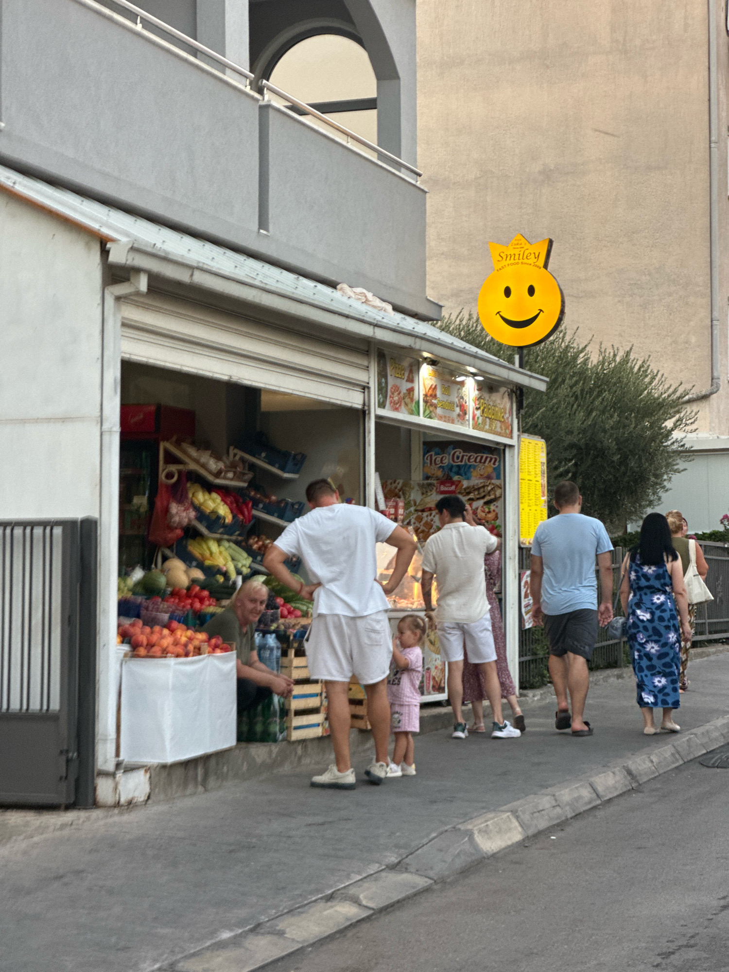 In Budva, Montenegro, on a street, a fruit stand is open for business. A man in a green shirt is attending to the fruits, which are displayed in crates and on tables. Piles of peaches are visible in the foreground. Several people are standing in front of the stand, some appearing to be customers. A man in a white t-shirt and white shorts is looking at the produce. A young girl in a pink outfit stands near him. Behind them, other individuals are walking past. A yellow sign with a smiley face and the text "Smiley Fast Food since 2009" is mounted on the wall of an adjacent building. The street appears to be paved, with a sidewalk in front of the stand. The buildings are typical of a Mediterranean urban environment.