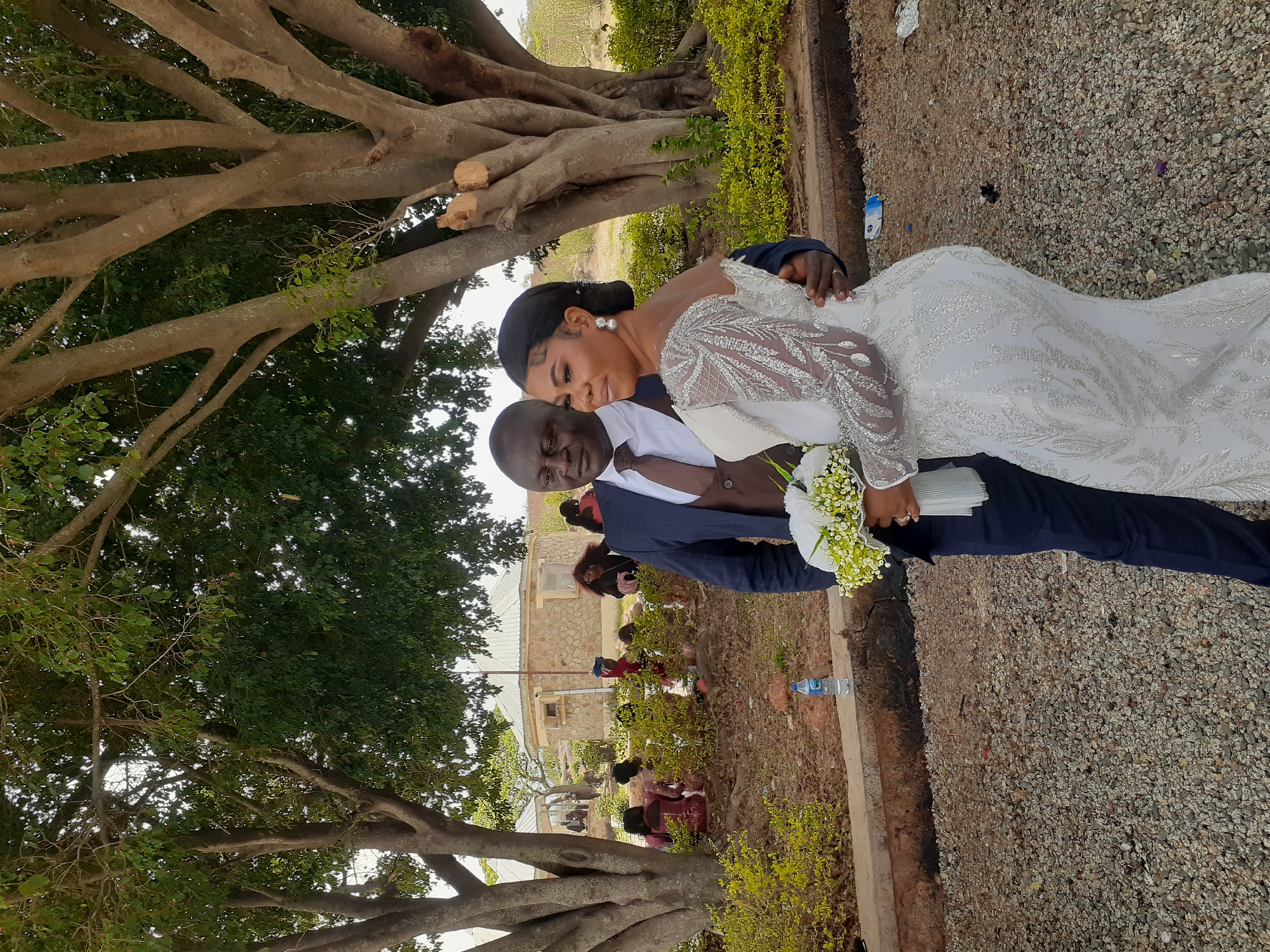 A newly married couple poses outdoors in Jos, Nigeria. The groom, dressed in a dark blue suit, holds a bouquet. The bride, wearing a white wedding dress with intricate detailing, embraces him. They stand on a gravel path bordered by a concrete curb and green foliage. A large, branching tree provides a backdrop. In the distance, a building and several people can be seen. A bottle of water rests on the ground near the curb.
