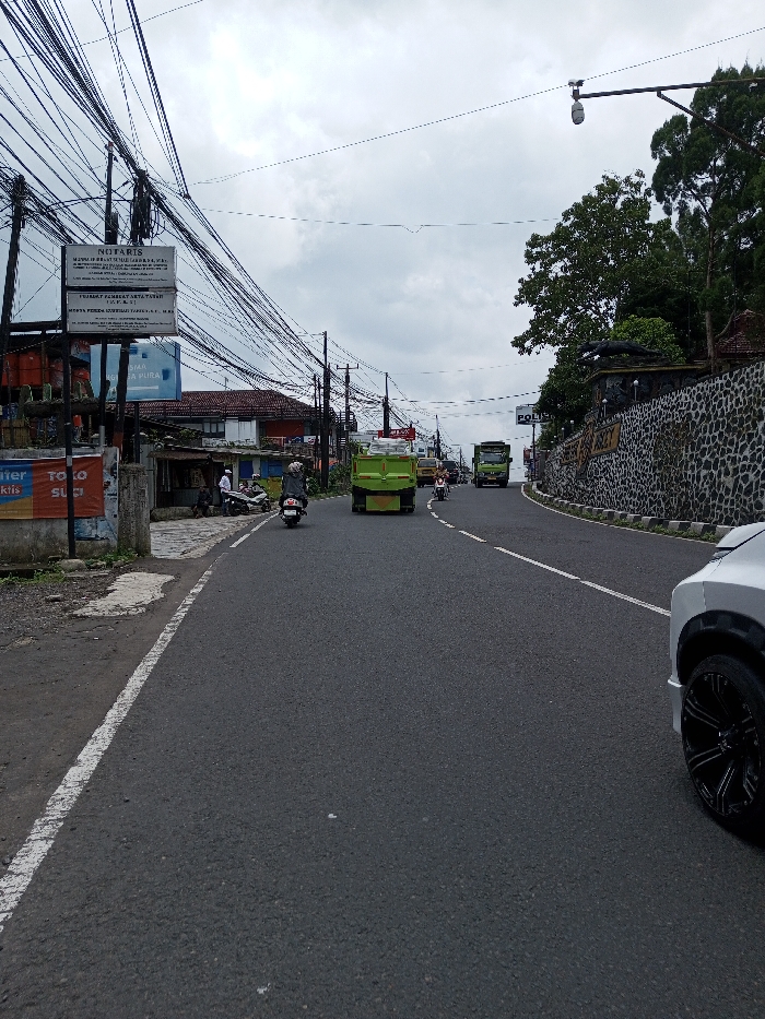 The scene is depicted in the city of Chendenbji, Bhutan. Under an overcast sky, an asphalt road with a white broken centerline curves slightly uphill. On the left, a series of utility poles supports a dense array of black electrical and communication cables. Attached to a pole are several signs: one reads 'NOTARIS,' another 'IJIN PRAKTIK NOTARIS,' and a blue sign partially displays 'PERURI' and 'PURA.' These visible texts are in Indonesian. Adjacent to the road, on the left shoulder, are low-rise buildings with tiled roofs and an open-front stall. Several individuals are present on the shoulder; one is near a parked scooter. Traffic on the road includes a green dump truck in the mid-ground, followed by a motorcyclist wearing a black jacket. Further ahead, another green truck and additional vehicles are visible. On the right side of the road, a textured stone retaining wall runs parallel to the asphalt. A sign on this wall is partially visible, displaying 'POLISI,' which is Indonesian for Police. Above the wall, substantial green trees and foliage are present, along with a dark, sculpted figure resembling an animal. A street light fixture extends over the road from a pole on the right. In the immediate foreground, the front fender and a black multi-spoke wheel of a silver vehicle are visible on the right.