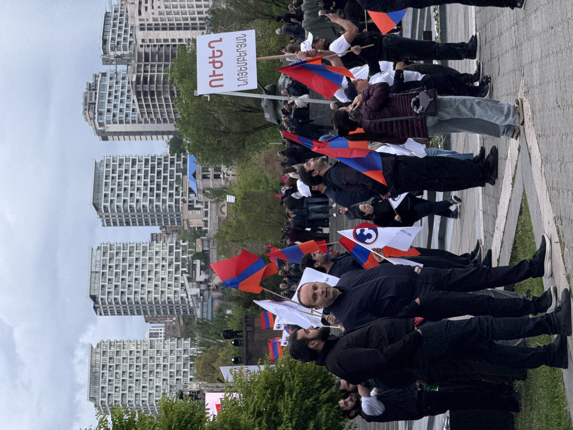 A crowd of people, many holding Armenian flags and signs with Armenian script, gather on a paved area in Yerevan, Armenia. Several tall, modern buildings stand in the background under a cloudy sky. The people are dressed in a variety of clothing and appear to be involved in a demonstration or rally.