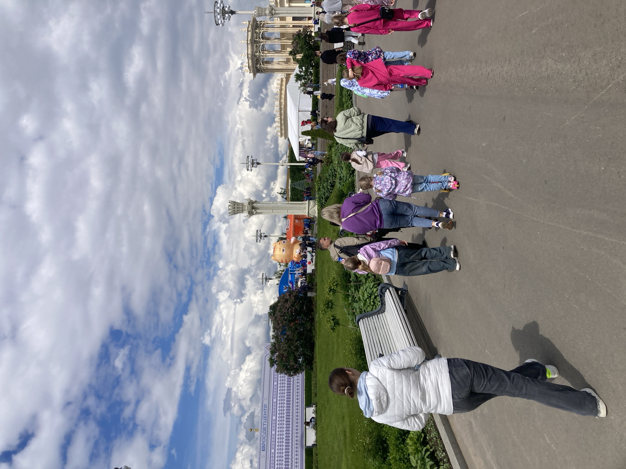 The image shows a scene in Moscow, Russia, likely at the Exhibition of Achievements of National Economy (VDNKh). The sky is cloudy with patches of blue. A group of people is walking on a paved path, including adults and children. In the foreground, a person in a white jacket is sitting on a park bench. A sign with text in Russian is visible. In the background, there are buildings and other structures typical of VDNKh, including lamp posts and pavilions. A large, inflatable cartoon character is also visible.