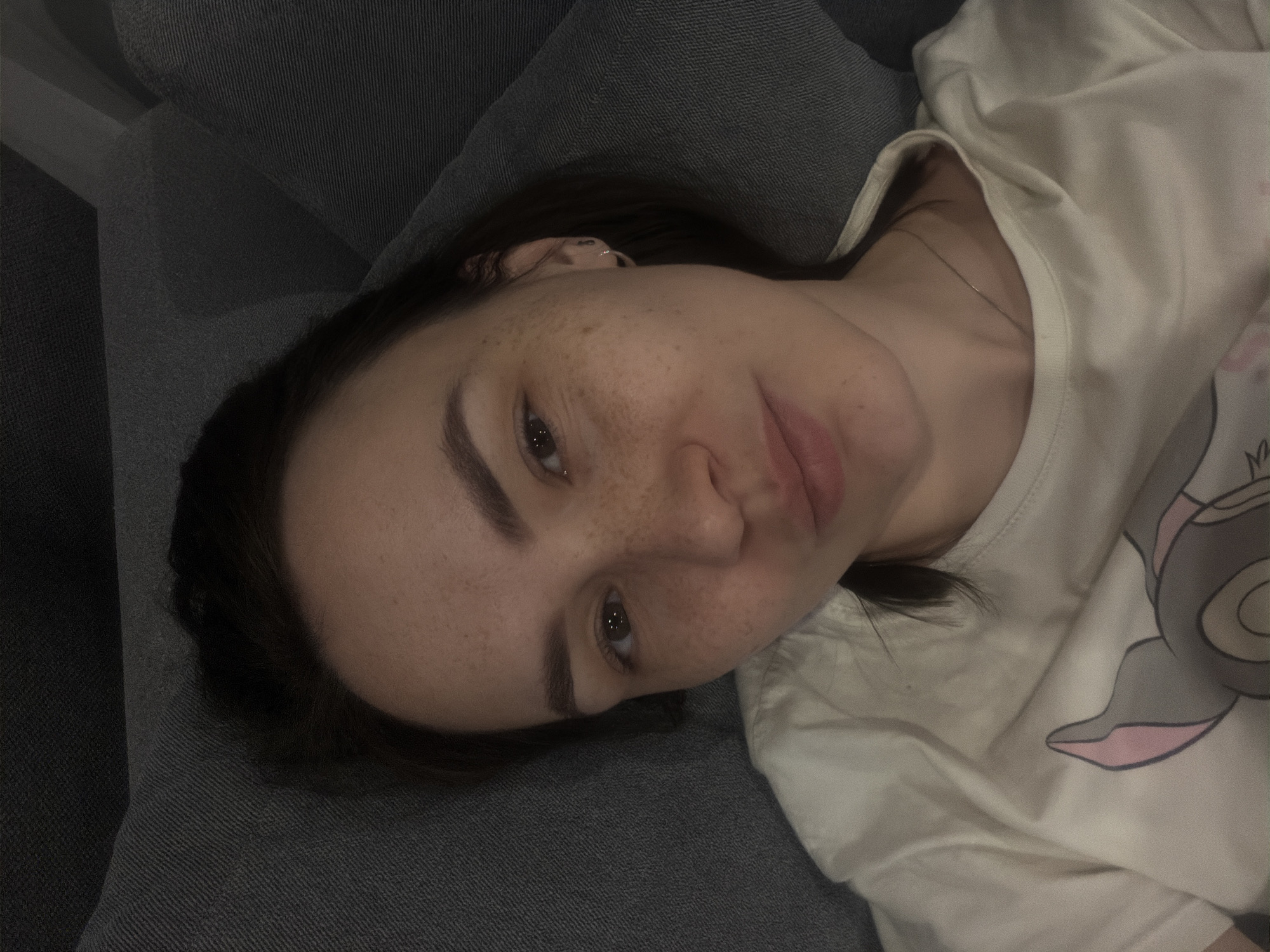 A person is lying on a gray upholstered surface, possibly a couch or bed. The person has fair skin, dark hair, and freckles. They are wearing a light-colored t-shirt with a cartoon-like image on it. The exact location in the city of Chaikovsky, Russia cannot be determined from the image.