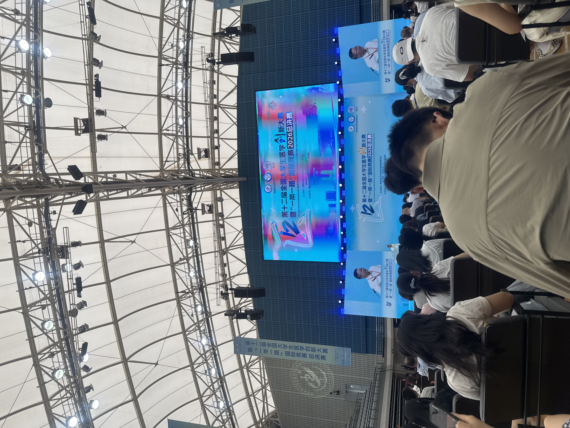 The image depicts a large indoor event, likely a conference or competition, with a sizable audience seated in rows of chairs facing a large screen. The screen displays text and graphics related to an event titled "The 12th China Undergraduate Medical Students' Innovation & Entrepreneurship Competition." Two individuals are visible on smaller screens within the main display, one speaking at a podium. The ceiling is a white, tent-like structure supported by metal beams and industrial lighting fixtures are suspended from above. Several banners are hanging from the ceiling, one featuring Chinese characters and the event title. The audience members are diverse in age and gender, with most facing the screen. The overall impression is of a formal, organized event taking place in a large venue. The specific location of Yangpu District, China is not directly identifiable from visual cues within the image.