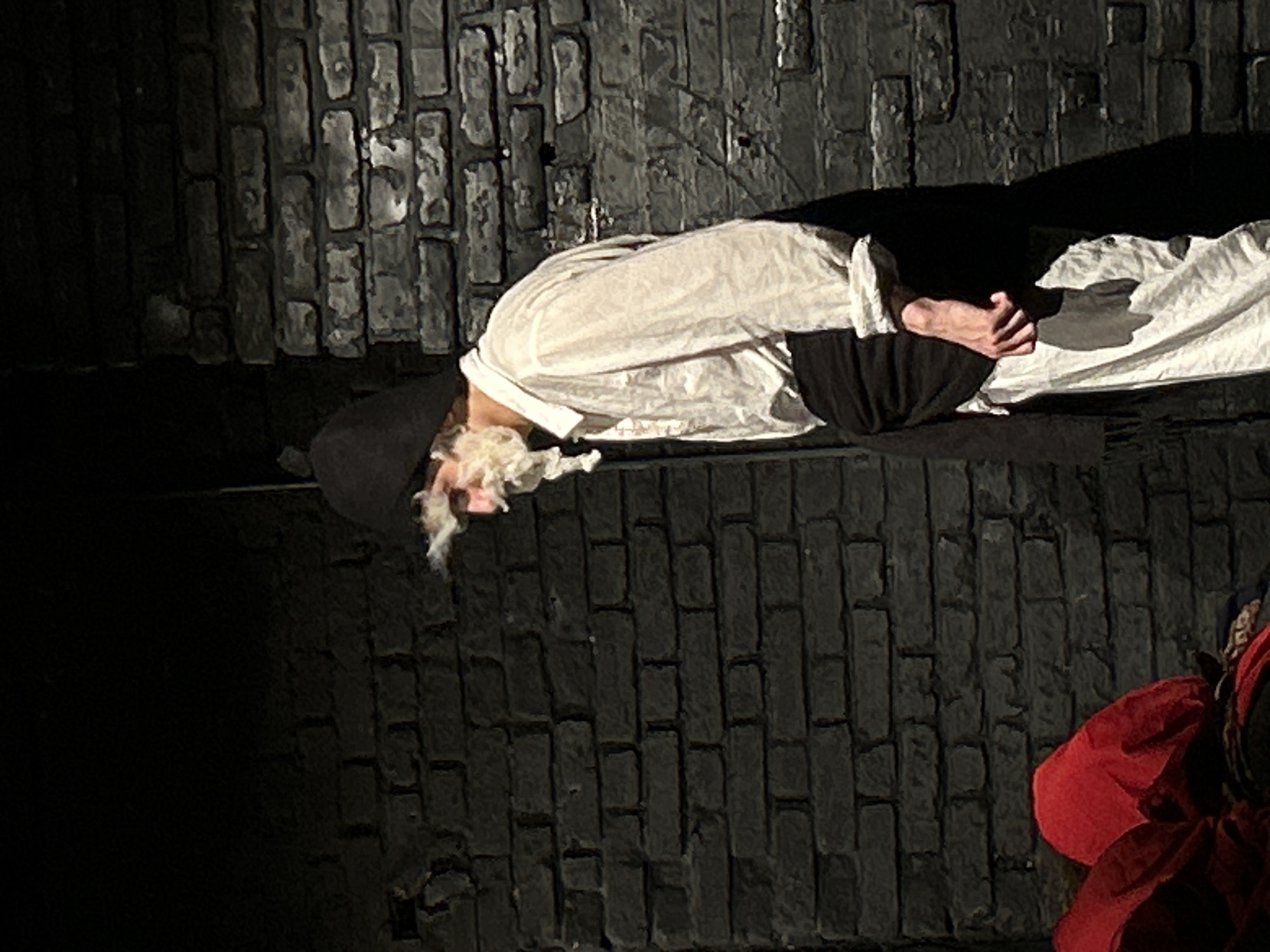 A figure in theatrical costume lies on a brick surface, presumably a stage, in a dimly lit environment. The figure, dressed in a white shirt or robe with black sleeves, wears a black hood obscuring their head and a white, disheveled beard or wig. The person's hands are clasped near their chest. The brick surface is dark and textured, with uneven lighting highlighting the individual bricks. At the bottom right corner of the image, a red garment is visible, hinting at the presence of another person or prop. The location appears to be within a theater or performance space.