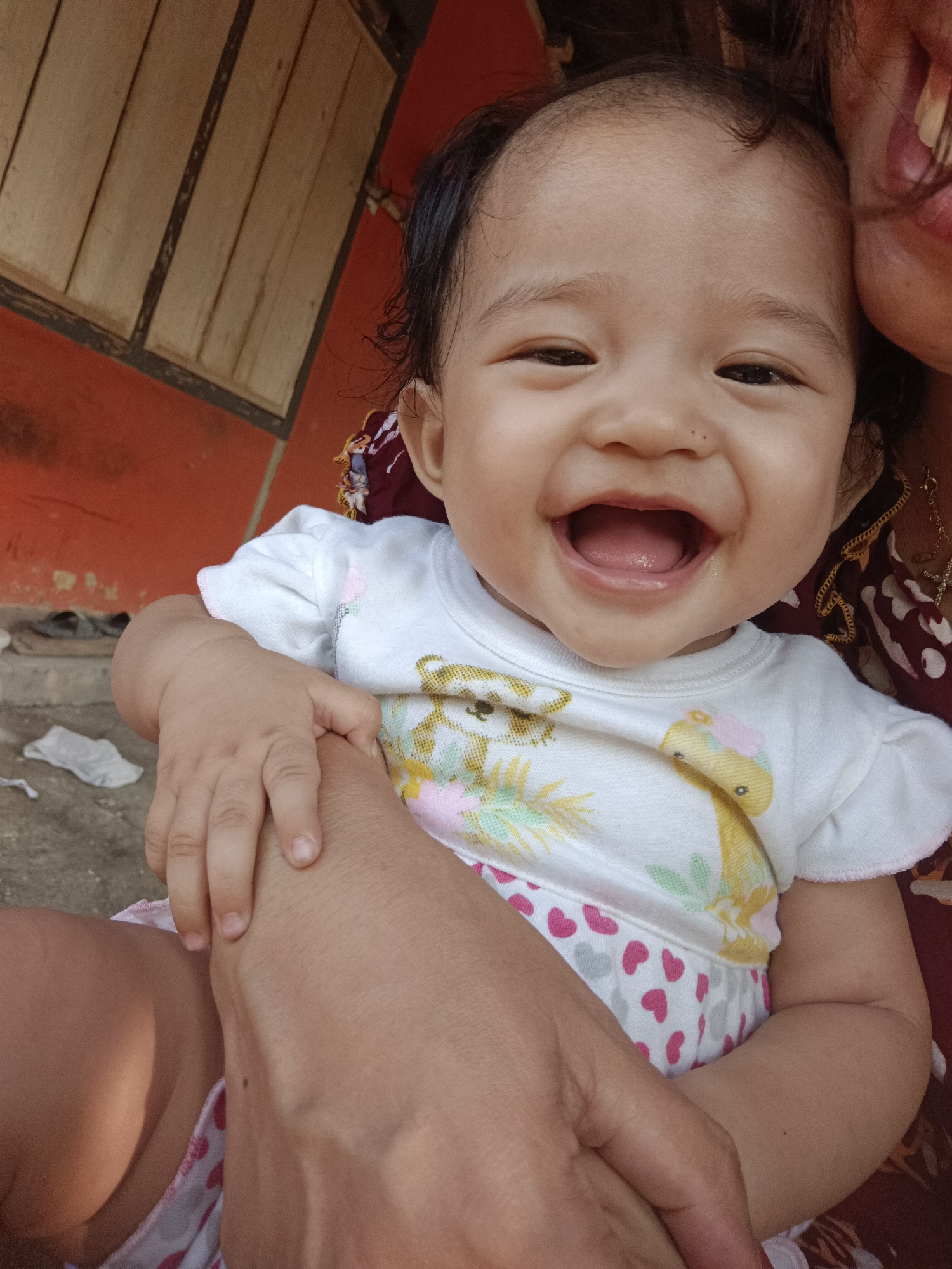 A close-up, high-angle medium shot captures a smiling infant with dark hair, looking directly at the camera in the city of Kawua, Indonesia. The infant is wearing a white t-shirt with a cartoon animal print and pink and white heart-patterned shorts. An adult's arm is visible cradling the infant. The background features a rustic wooden structure with orange walls and a concrete floor. The lighting is bright, suggesting daylight conditions.