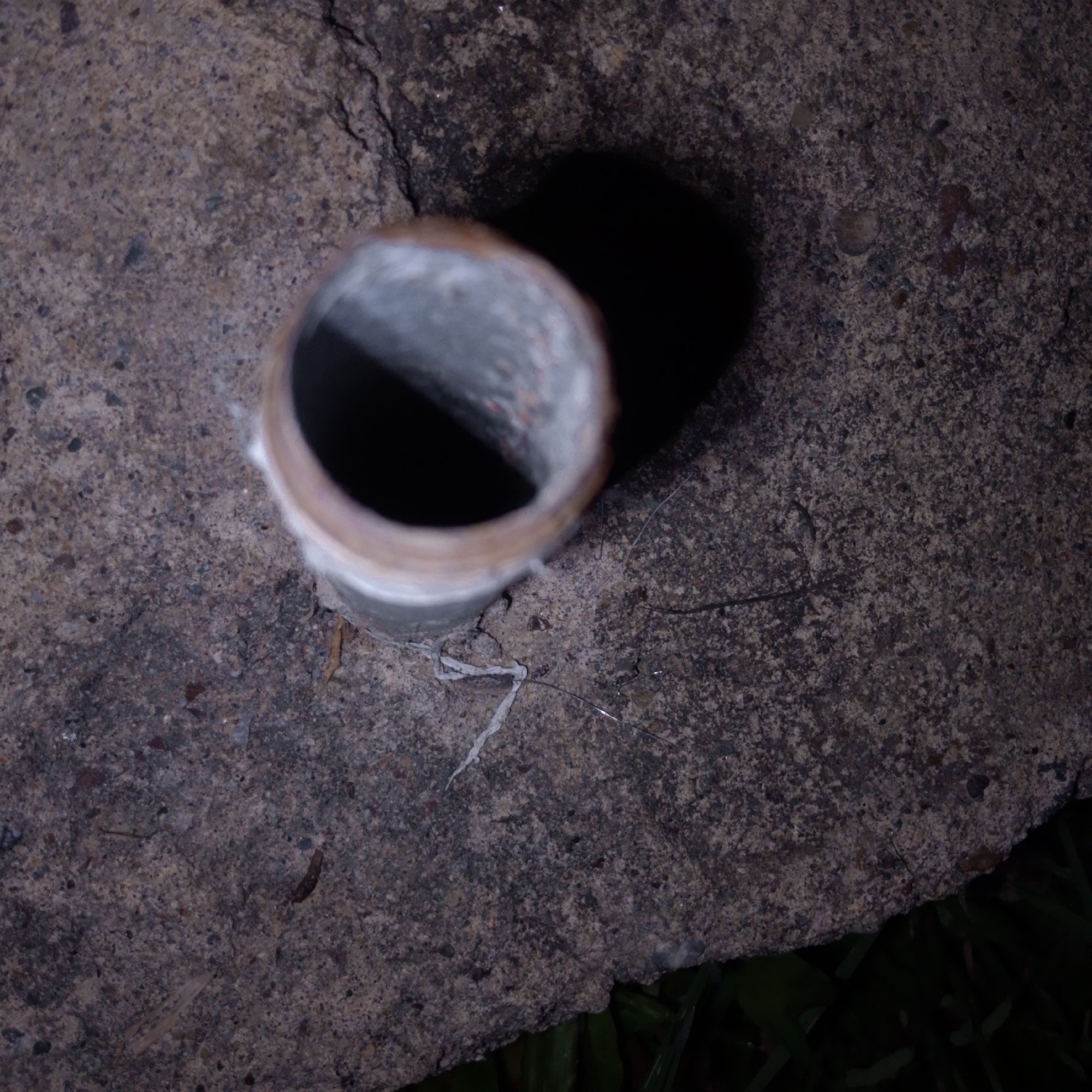An overhead shot shows a metal pipe protruding from a concrete surface. The pipe is cylindrical, with a textured interior, and appears to be rusted or discolored on the exterior rim. A dark shadow is cast to the right of the pipe. The concrete surface is rough and textured, with some cracks and debris. Fragments of white material, possibly plastic, are near the base of the pipe. Patches of dark green vegetation are visible in the lower right corner. No information about the location in Lhokseumawe, Indonesia is visible.