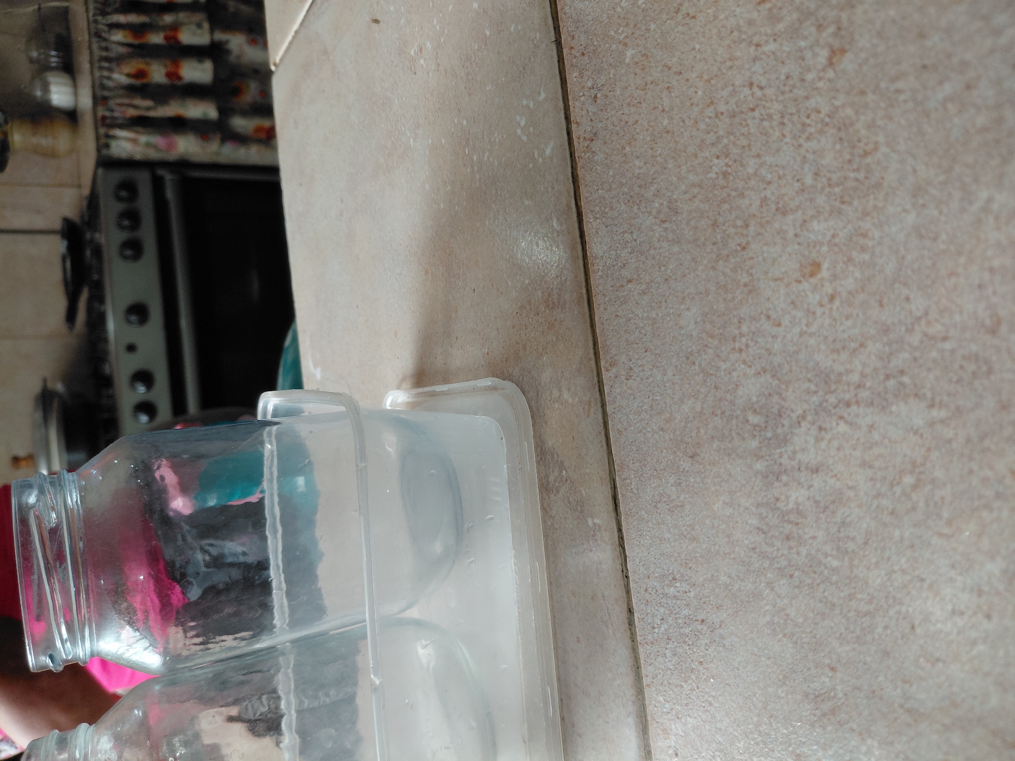 The image, oriented on its side, provides a close-up view of a domestic kitchen setting. In the foreground, two clean, empty glass jars are placed within a clear plastic container, which appears to be a draining rack or storage tray as it holds some water residue. These items rest on a light-colored, possibly beige or tan, tiled countertop featuring a speckled pattern and a visible grout line.
In the blurred background, to the left of the counter, a part of a kitchen appliance, likely a stove or oven, is visible with several circular control knobs. Above the appliance, there is a patterned surface, which could be a wall or backsplash, exhibiting dark and reddish-brown textures. A reflection or view through one of the glass jars reveals a hint of pink or red fabric, possibly from a person's clothing. The overall scene suggests items are being dried or stored in a kitchen during the daytime. No text is visible in the image.