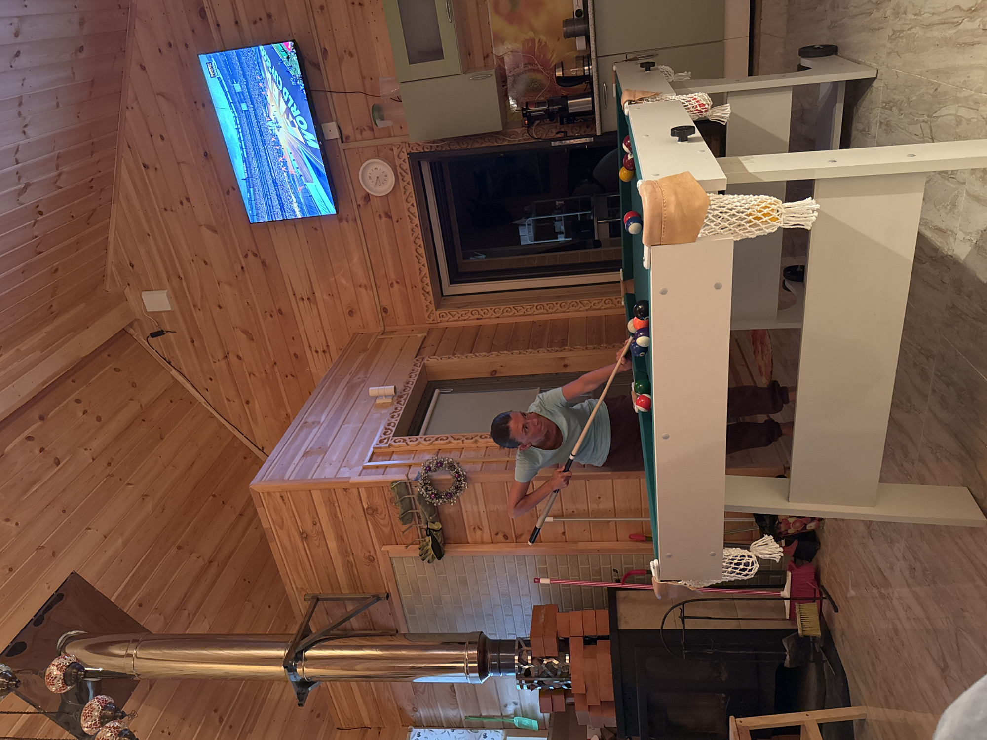 The image shows a woman playing billiards in a room with wood-paneled walls and ceiling. The room appears to be in a home or a recreational facility. A large metal chimney is visible on the left side of the image, along with a fireplace. A television is mounted on the wall, displaying what appears to be a sporting event. A clock is also visible on the wall. The pool table has several balls on it, and the woman is in the process of taking a shot. The overall atmosphere is casual and relaxed. This image does not contain any information that would allow for the localization to Zelenaya Roshcha, Russia.