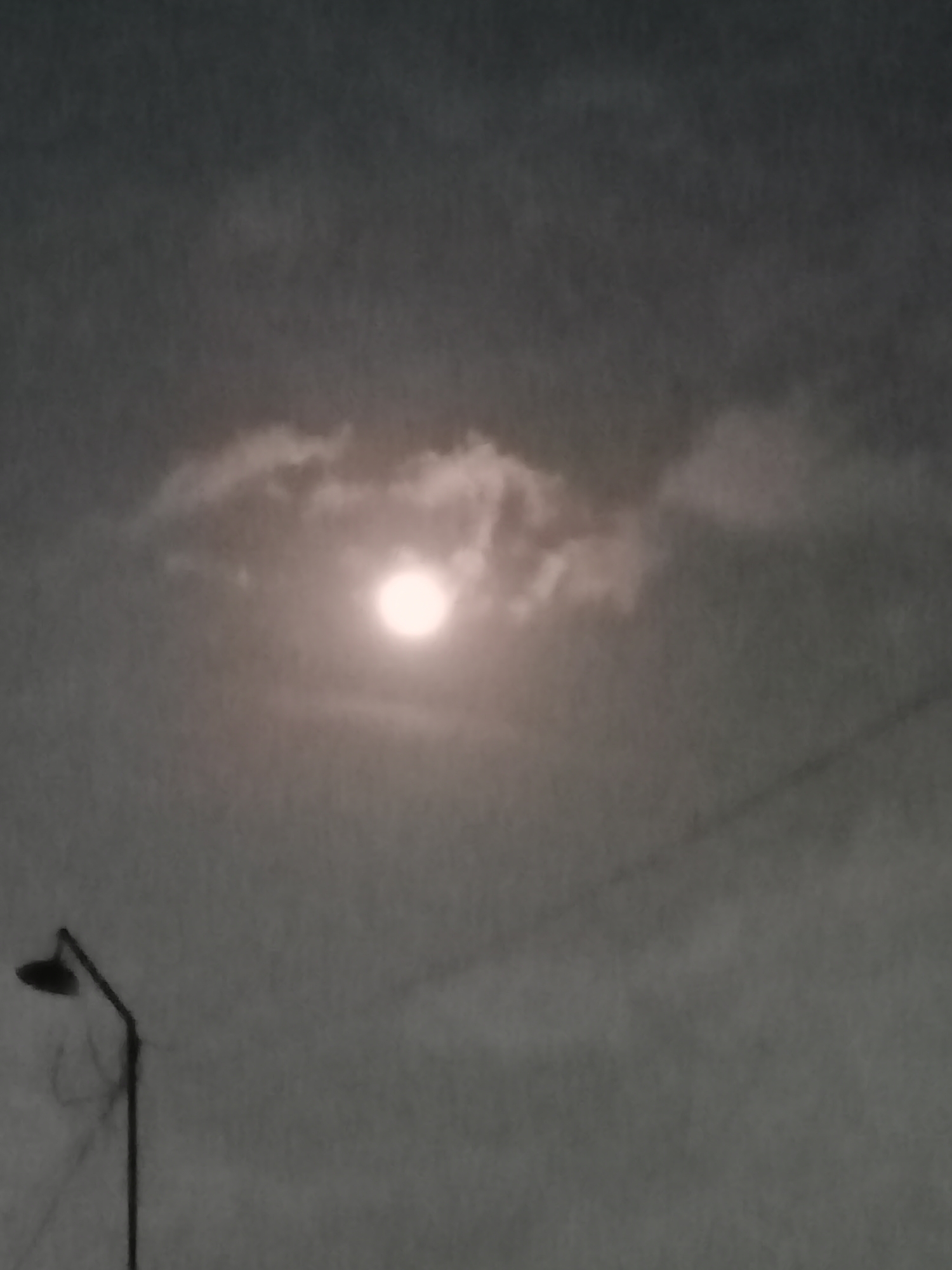 The image shows a hazy, overcast sky with a bright, diffused circle, likely the moon, partially obscured by clouds. In the lower left quadrant, a dark silhouette of a street lamp is visible, with its pole extending upwards. The overall mood is dim and atmospheric, suggesting it was taken at night or during twilight. There are no people or discernible structures beyond the lamp post.