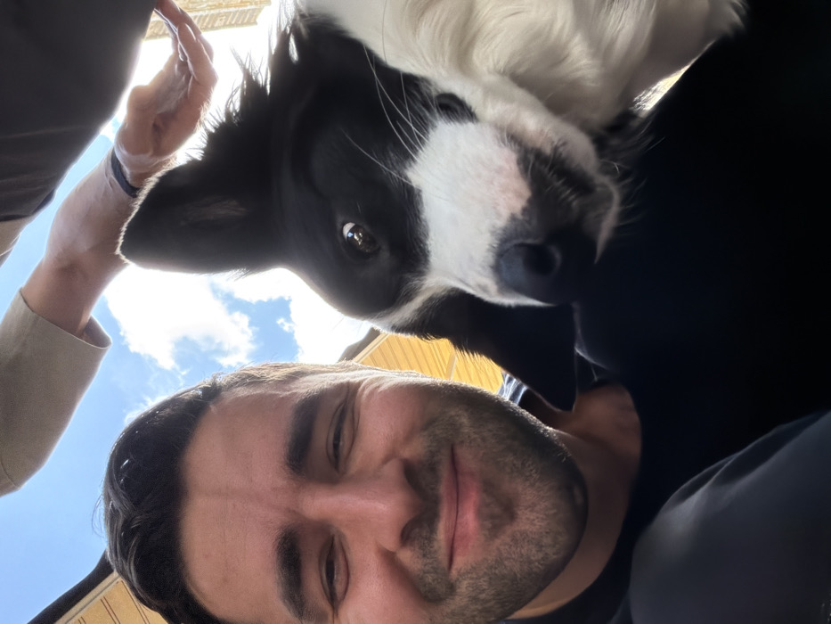 An adult male occupies the left foreground, visible from the forehead to mid-chest. He has dark hair, facial stubble, and a smiling expression directed towards the camera. He wears a dark t-shirt. To his immediate right, a black and white dog, consistent with a Border Collie, is positioned with its head tilted. The dog displays one erect ear and one folded ear; its left eye (viewer's right) is open and exhibits a light reflection. A human forearm and open hand extend from the upper right, positioned above the dog, implying an interaction such as petting. A dark wristband is present on the wrist, and the arm is associated with a light-colored garment. The background features a bright blue sky with scattered white clouds, indicating an outdoor, daytime setting. A blurry, light-colored structure with vertical lines, possibly an awning or building facade, is discernible in the upper left background. The scene is situated in Tel-Aviv, Israel.