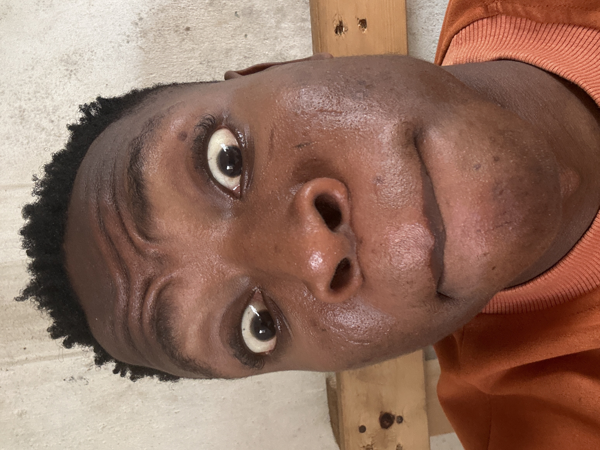 The media file shows a close-up, overhead shot of a person's face. The person appears to be young, with dark skin and short, curly hair. They are wearing an orange sweatshirt. Their eyes are open wide, and they have a slight smile on their face. The background is out of focus but appears to be a wall and some wooden beams, suggesting an indoor or possibly sheltered outdoor setting. The lighting is warm and even, indicating it's likely daytime. The image has a casual, selfie-like feel, and the person's expression could be interpreted as surprised or amused. No text is visible in the image. Based on the provided location context of Ikorodu, Nigeria, the scene likely takes place in this region.