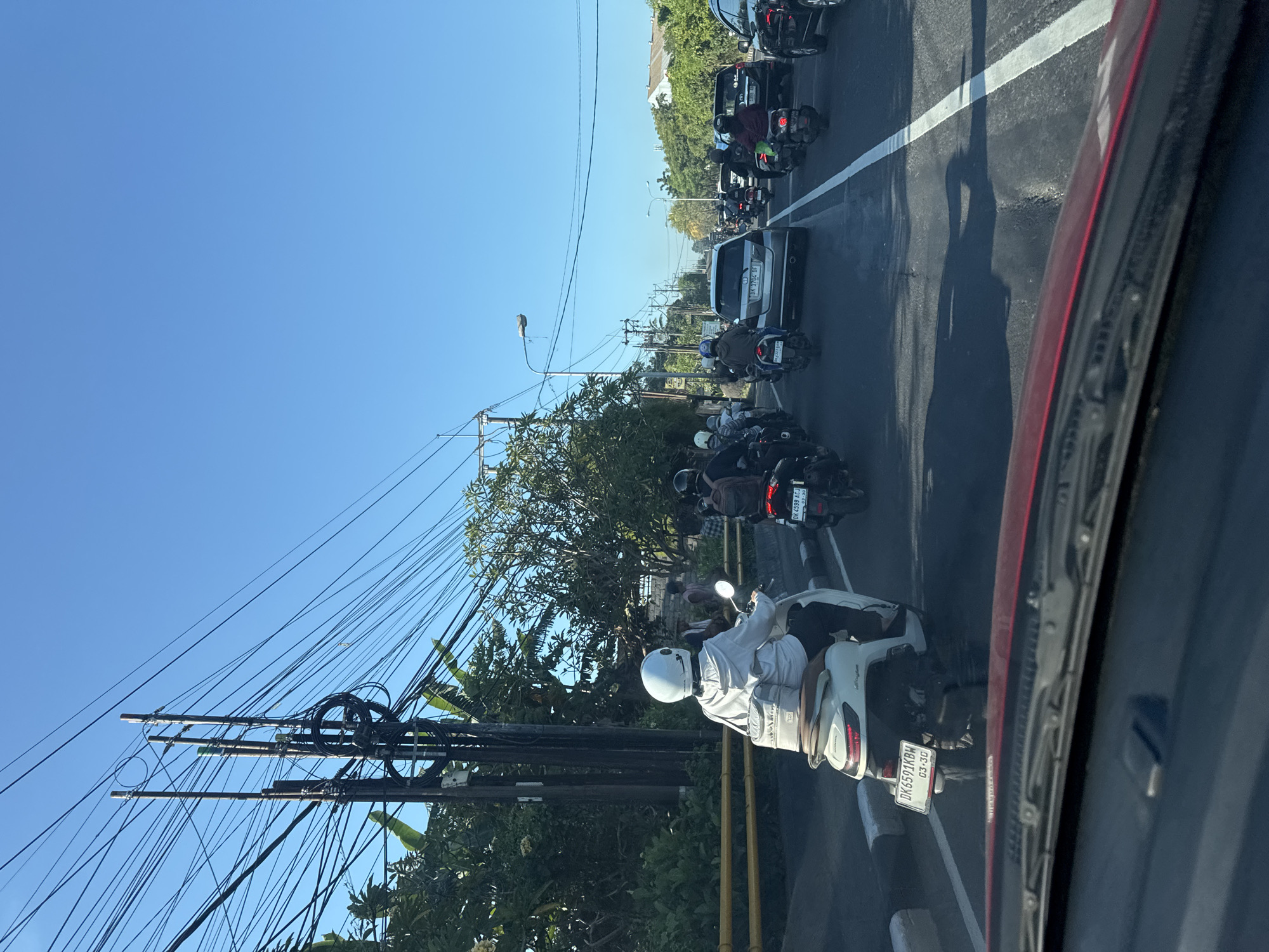 The image shows a congested road in Denpasar, Indonesia, with heavy traffic of motorcycles and cars. The viewpoint is from inside a car, looking towards the front. Numerous power lines run along the road, and trees line the sides. The sky is clear and blue. Motorcyclists are wearing helmets and are navigating the traffic. The license plates of the vehicles are visible. The dense traffic is typical of urban areas in Bali.