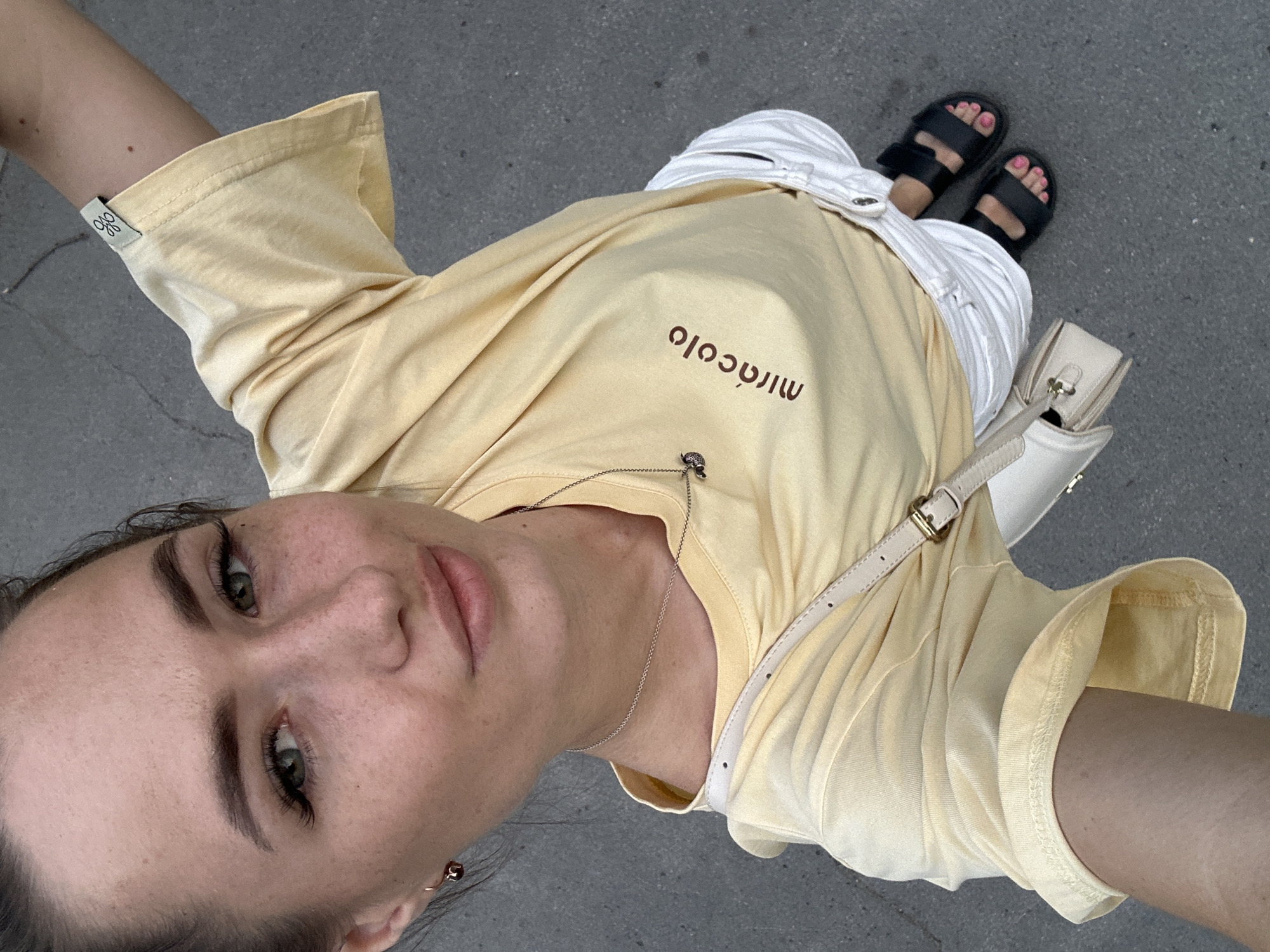 A woman is standing on a paved surface, possibly a street or sidewalk, and taking a selfie from a high angle. She has light skin, green eyes, and dark hair pulled back. She is wearing a yellow t-shirt with the word "miracolo" printed on it in brown letters, white shorts, and black sandals. She has a small, light-colored crossbody bag slung over her shoulder and is wearing a delicate silver necklace with a small pendant. The background is a gray, cracked pavement.