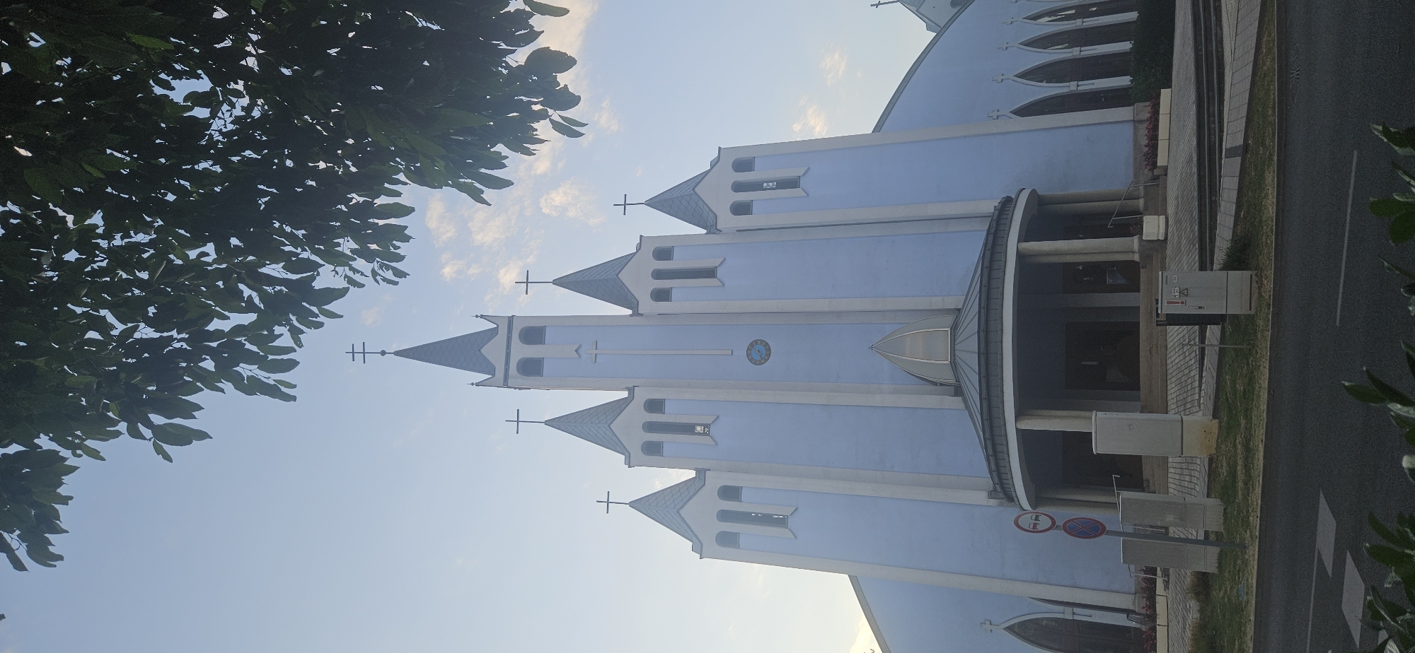 This is a vertical, eye-level shot of the exterior of a pale blue church in Hévíz, Hungary. The church features a symmetrical facade with multiple towers topped with crosses. The central tower is the tallest and has a cross at its apex. There are five towers visible in total, with three more on the right side extending further back. The church has a rounded, covered entryway with columns. A clock face is visible on the central part of the church. The sky is a light blue with some wispy white clouds. The top of the image is partially obscured by dark green leaves from a tree. In the foreground, there is a paved area with some concrete utility boxes and traffic signs. A road is visible in the lower part of the image, with some greenery in the very bottom. No people are visible in the frame.
