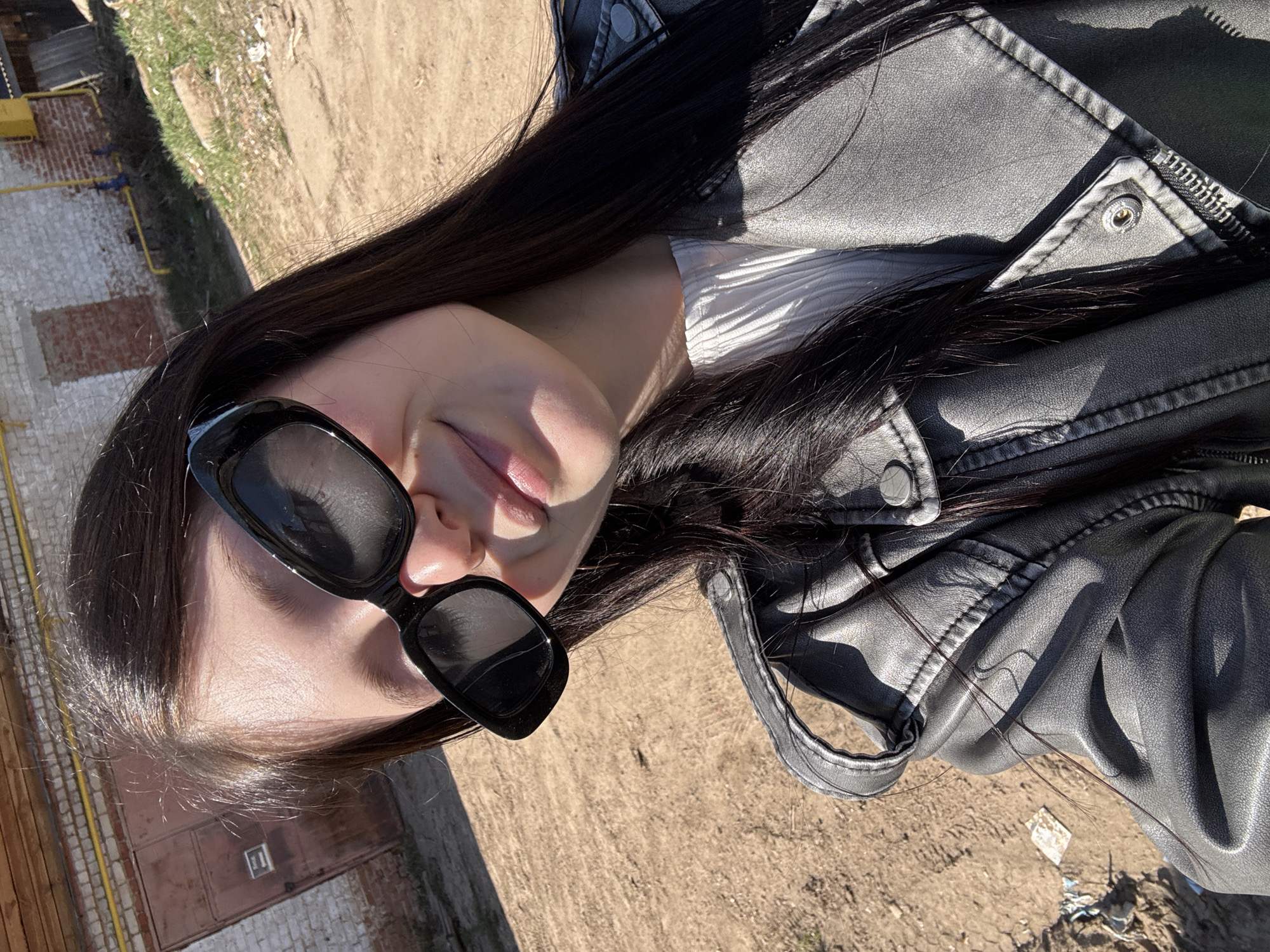 The image shows a woman taking a selfie outdoors in the city of Фоки, Russia. She is wearing sunglasses and a black leather jacket over a white top. The background appears to be a dusty outdoor area, possibly an alley or side street, with a brick structure visible in the corner of the frame. The lighting suggests it is daytime.
