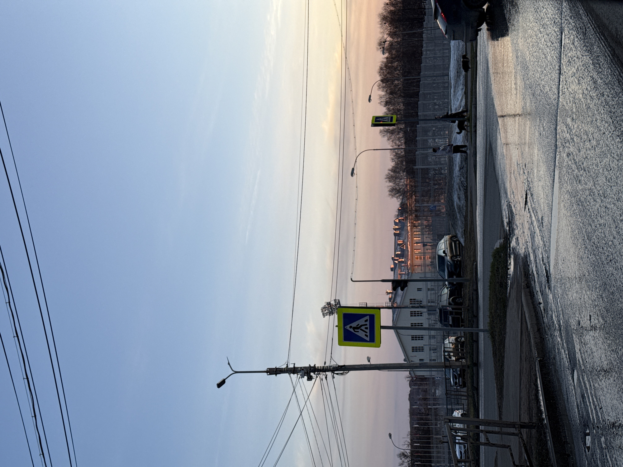 The image captures a street scene in Votkinsk, Russia, during twilight. The road is wet, reflecting the sky's soft colors. A pedestrian crossing sign stands prominently in the foreground. The sky is a gradient of pale blue to soft pink. Buildings are visible in the background. Power lines traverse the upper left portion of the frame. In the background, figures are seen crossing the street. Several parked cars line the roadside.