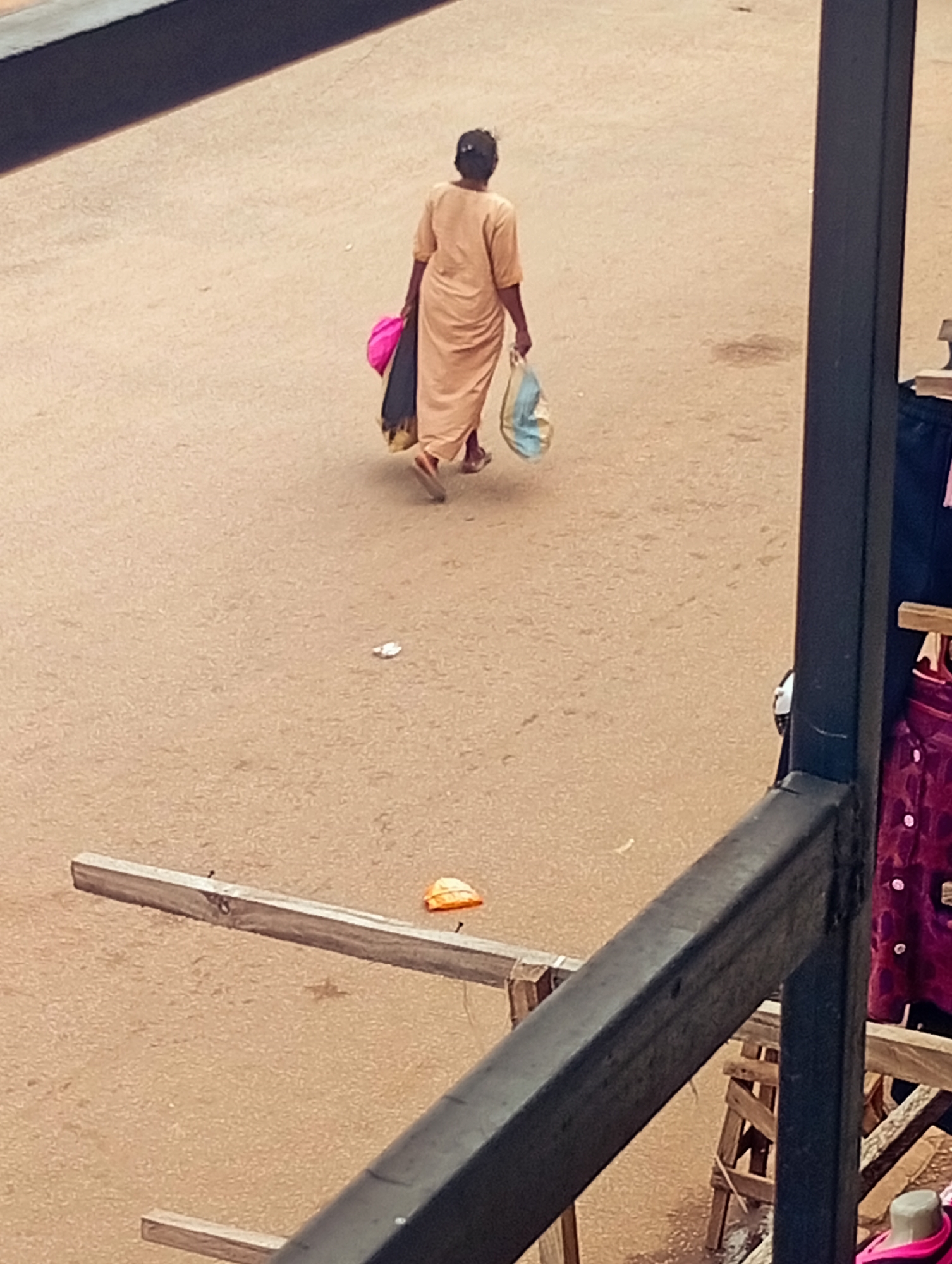 In Bukuru, Nigeria, a lone individual walks away from the camera on a dusty, light brown street. They are wearing a long, light orange dress and carrying two bags. One bag, held in their right hand, is a light blue and white patterned shopping bag. The other, in their left hand, appears to be a bright pink bag with dark fabric spilling out. The individual is walking towards the right side of the frame. In the foreground, a dark, thick railing is visible, obscuring the lower part of the image. To the right, a glimpse of a market stall is present, with a burgundy-colored garment displayed, adorned with white buttons. Scattered on the street are a few small, indistinguishable objects, including what appears to be an orange item near a piece of weathered wood in the mid-ground. The lighting suggests it is daytime.