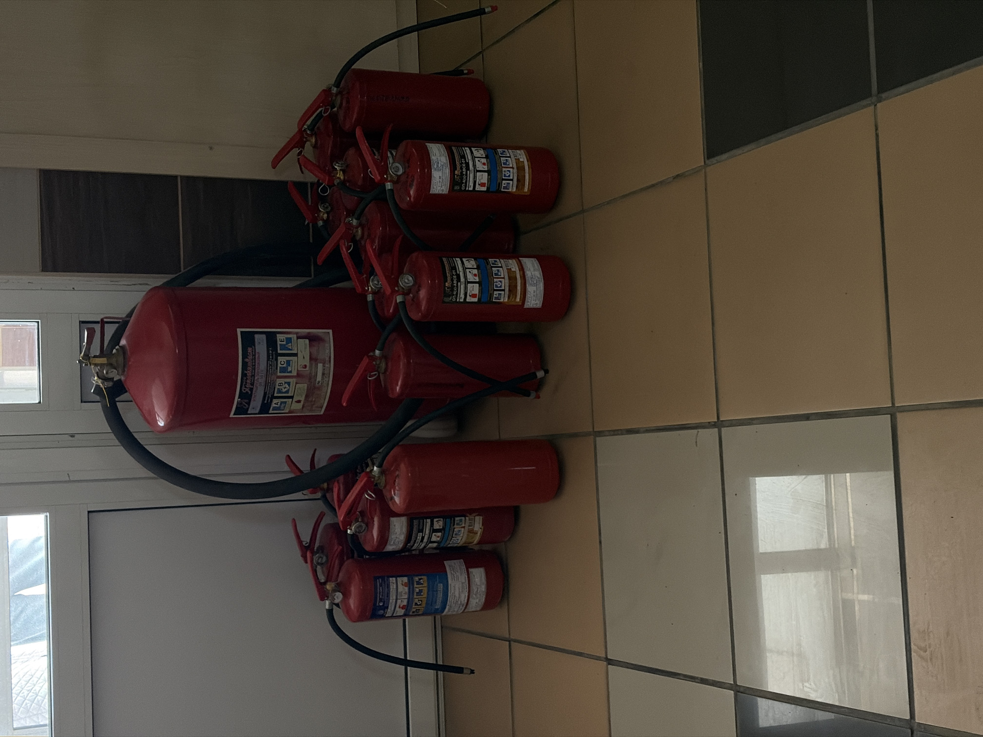 The image shows a collection of fire extinguishers in what appears to be an indoor environment. There's a large fire extinguisher attached with a hose to a series of smaller ones. The extinguishers are red with black hoses and various labels in Russian. The background includes a wall with tile and a window frame on the left side. The labels on the fire extinguishers appear to say "Приобинвес" (Priobinvest), “ОП-4(3)-АВСЕ-01” (OP-4(3)-ABCE-01), a fire extinguisher type. The exact location is not evident in the image beyond the fact that this brand is Russian. Therefore, it is not possible to conclusively determine that this image came from the city of Chaikovsky, Russia.