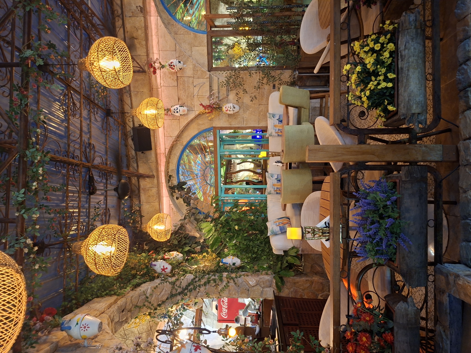 This image depicts an indoor/outdoor restaurant setting in Pattaya City, Thailand. The establishment features a blend of natural and artificial elements, including hanging wicker lamps, stone walls adorned with foliage, and decorative ceramic masks. Seating areas consist of wooden tables and cushioned chairs. Various plants, including yellow and purple flowers, are strategically placed around the area. In the background, a glimpse of the restaurant's interior reveals a Coca-Cola refrigerator. The setting suggests a combination of rustic and decorative aesthetics.