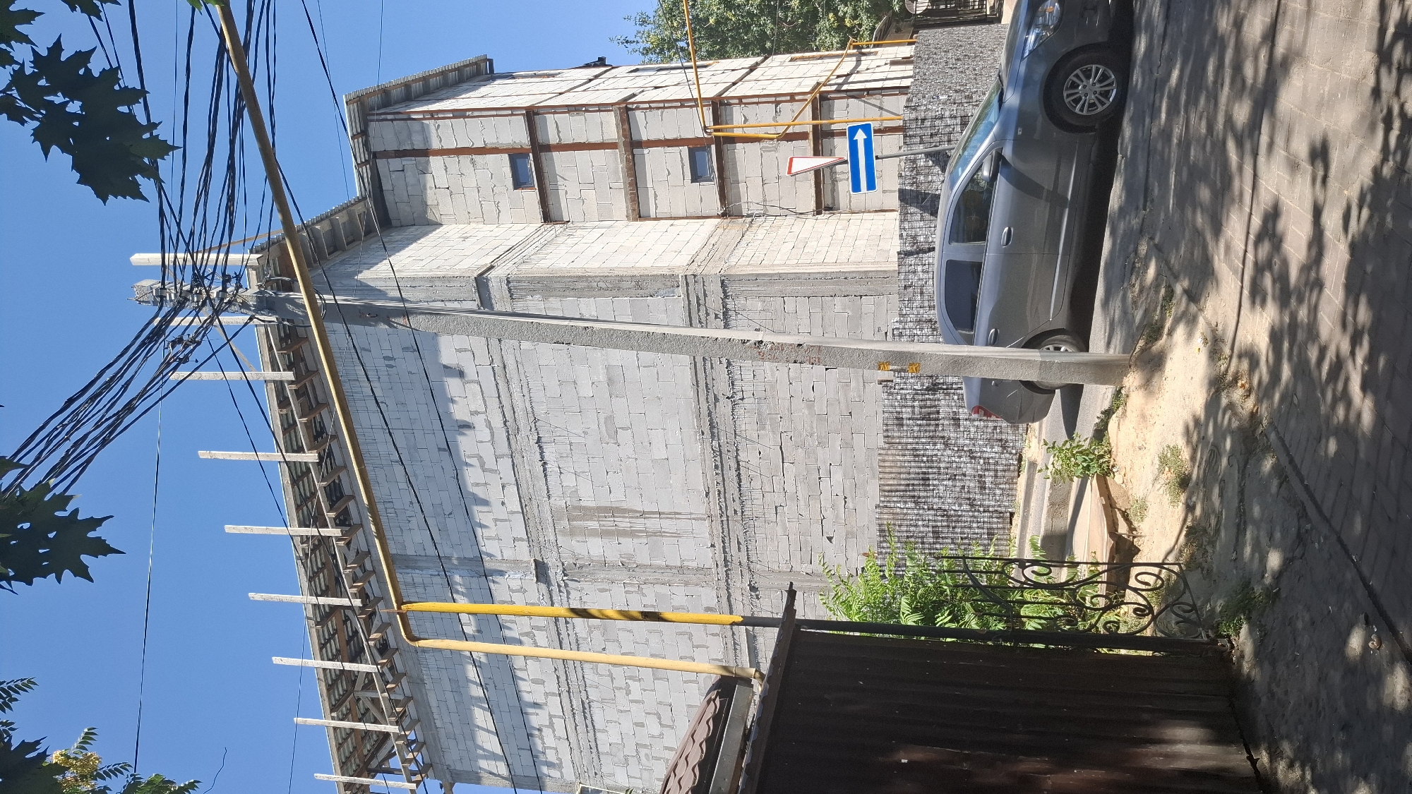 A grey sedan is parked on the right side of a paved street in front of a construction site. The construction site features a multi-story building made of cinder blocks with wooden beams extending from the roofline. A yellow pipe runs vertically along the side of the building, and a grey pole with wires stretches across the scene. A blue traffic sign with a white arrow indicating a right turn is mounted on a pole near the building. The ground in the foreground is a mix of pavement and sand, with some vegetation growing around a black metal gate on the left. The sky is clear and blue.