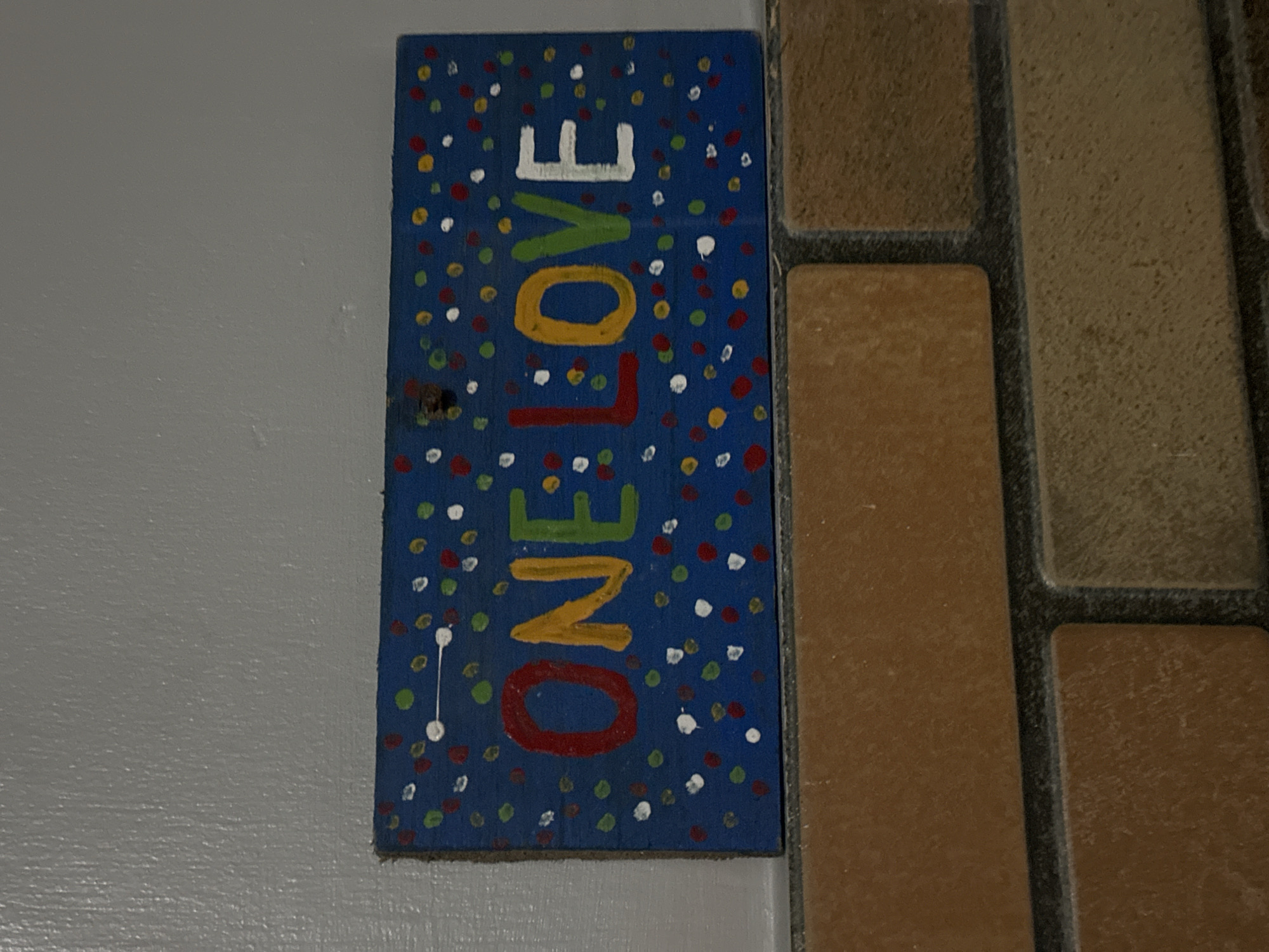 A sign reading "ONE LOVE" hangs on a wall. The sign is a rectangular piece of wood, painted blue with small dots of various colors scattered across the surface. The words "ONE LOVE" are painted vertically on the sign in different colors, with "O" in red, "N" in yellow, "E" in green, "L" in white, "O" in yellow, "V" in green, and "E" in white. The wall to the left of the sign is light gray. To the right of the sign are light brown bricks. The location is North Kuta, Indonesia.