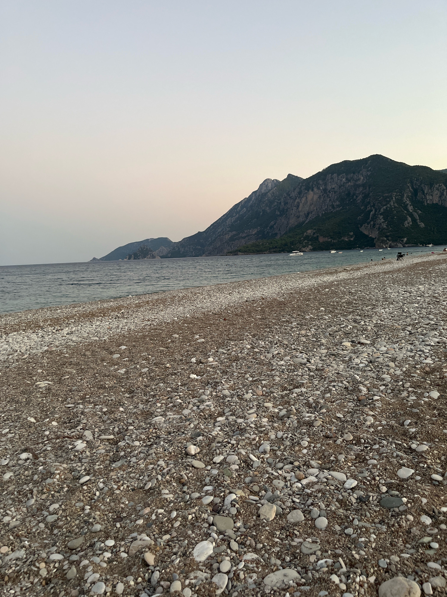 The image captures a wide, pebble-strewn beach in Kemer, Turkey, with the Mediterranean Sea in the background. In the distance, a rugged, tree-covered mountain range dominates the horizon under a pale, twilight sky. Several small boats are visible on the calm, rippling water, and a few people can be seen enjoying the beach, some swimming and others relaxing on lounge chairs. The foreground is filled with a dense texture of various-sized pebbles and stones.