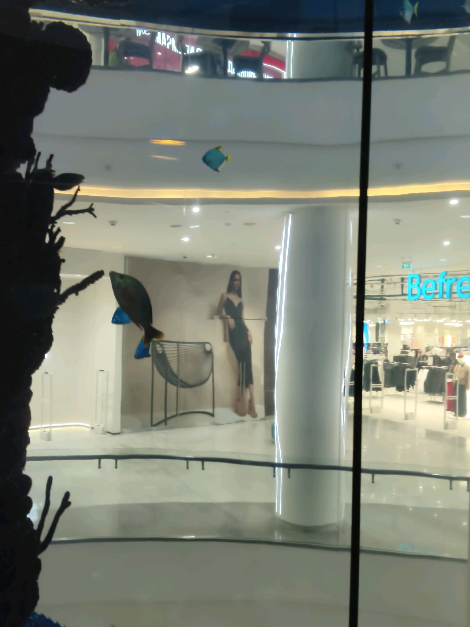 The image shows an indoor shopping mall. In the foreground, a large aquarium with a dark, shadowy decorative element resembling coral is visible. Two artificial fish, one blue and one with a dark body and orange tail, are suspended in the water. In the background, there is a large poster of a woman in a black dress, a seating area with a modern chair, and a store with the word "BeFree" visible on its signage. The mall appears to be multi-level, with shops and railings visible on an upper floor. The overall lighting suggests it is daytime, with artificial lights illuminating the interior. The presence of the "BeFree" store, which is a Russian clothing retailer, indicates the location is likely in Russia. Given the mall setting and the visible signage, it is plausible this image was taken in Moscow, Russia.