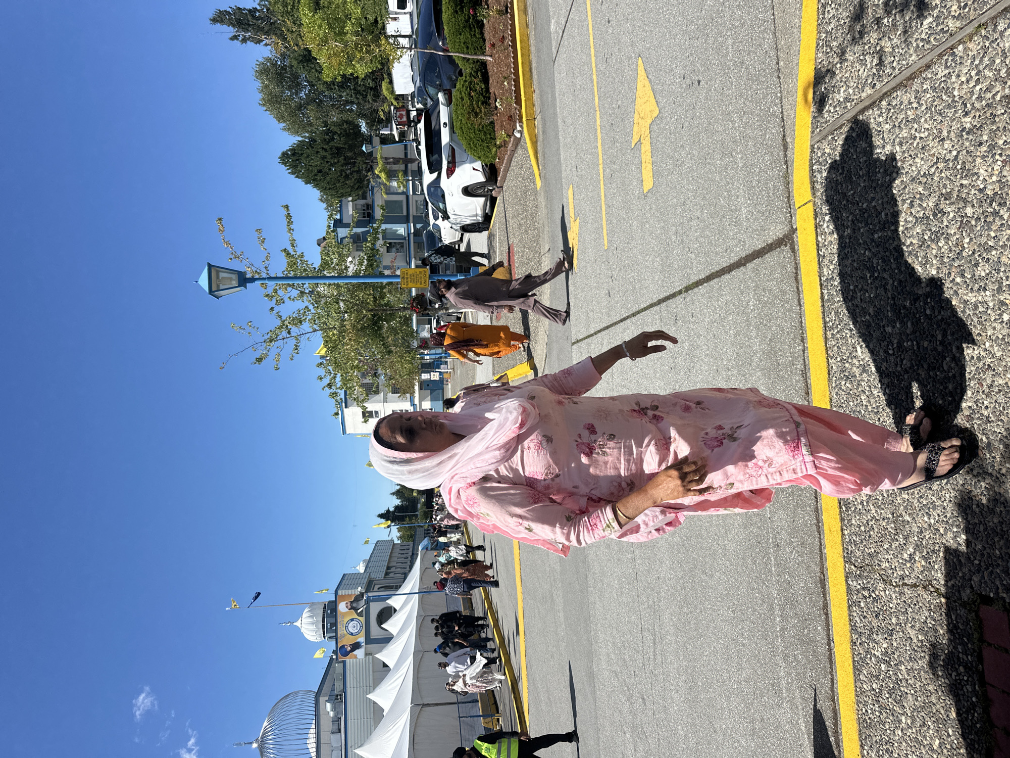 The image depicts a sunny outdoor scene outside a Sikh temple, likely the Gurdwara Sahib Sikh Society of Surrey, located in Surrey, Canada. In the foreground, a woman is walking to the right, wearing a pink floral salwar kameez and a light pink headscarf. She is wearing black sandals. Behind her, other people are walking in the same direction, some wearing traditional Sikh attire. The temple building, with its white domes and blue accents, is visible in the background. Tents are set up in front of the temple, and cars are parked in a lot to the right. Yellow lines are painted on the asphalt road, and a blue lamppost stands on the right side of the image. The sky is clear and blue.