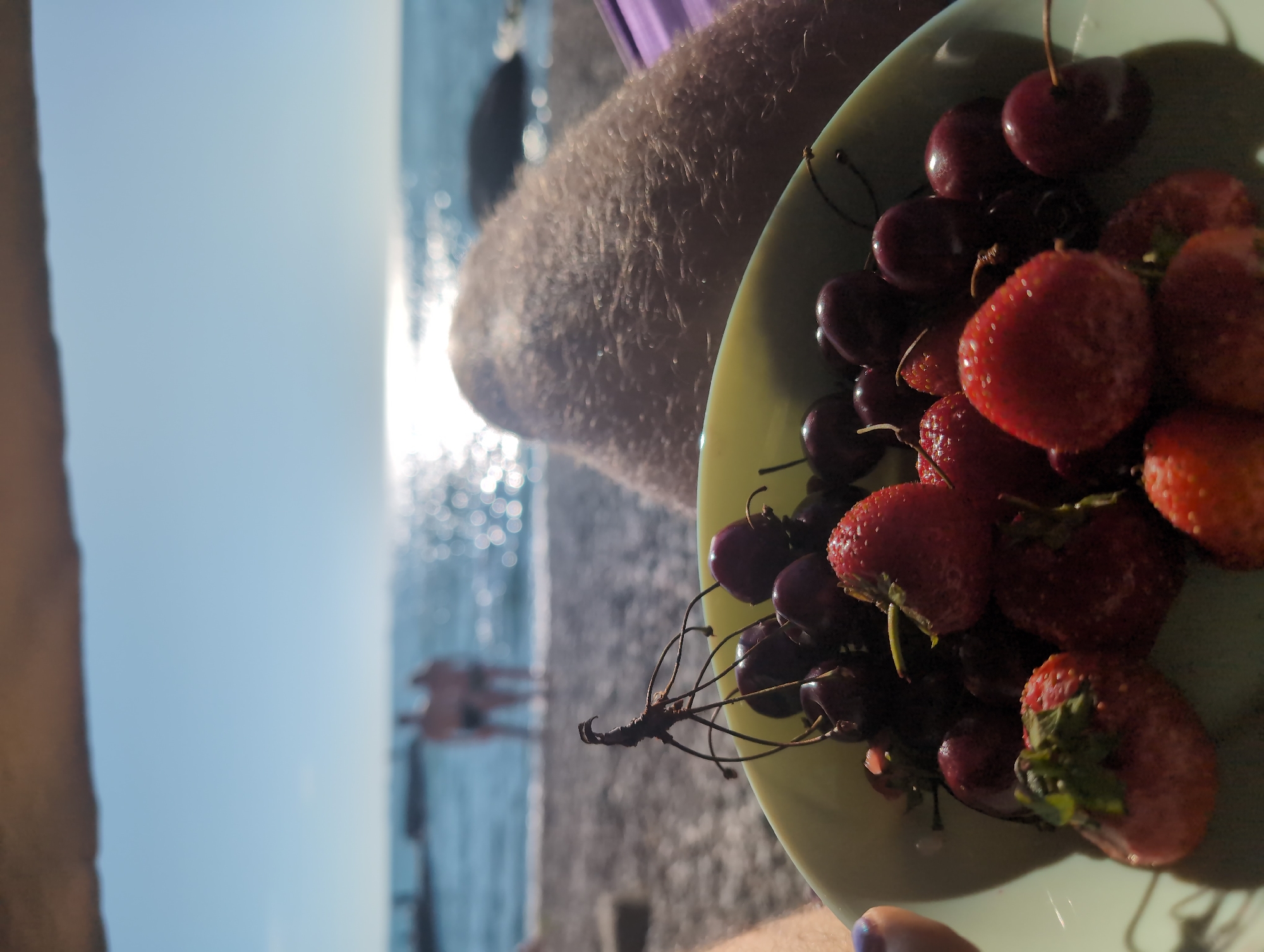 In Odesa, Ukraine, at the beach, a light-colored bowl overflows with ripe strawberries and cherries. The fruits are the focal point, with the strawberries displaying their characteristic red hue and textured surfaces, while the cherries are a deep, glossy red. In the background, the Black Sea glitters under a bright sun, its surface reflecting shimmering light. Two figures, indistinct and blurred, stand near the water's edge. The overall scene conveys a sense of relaxation and summer enjoyment.