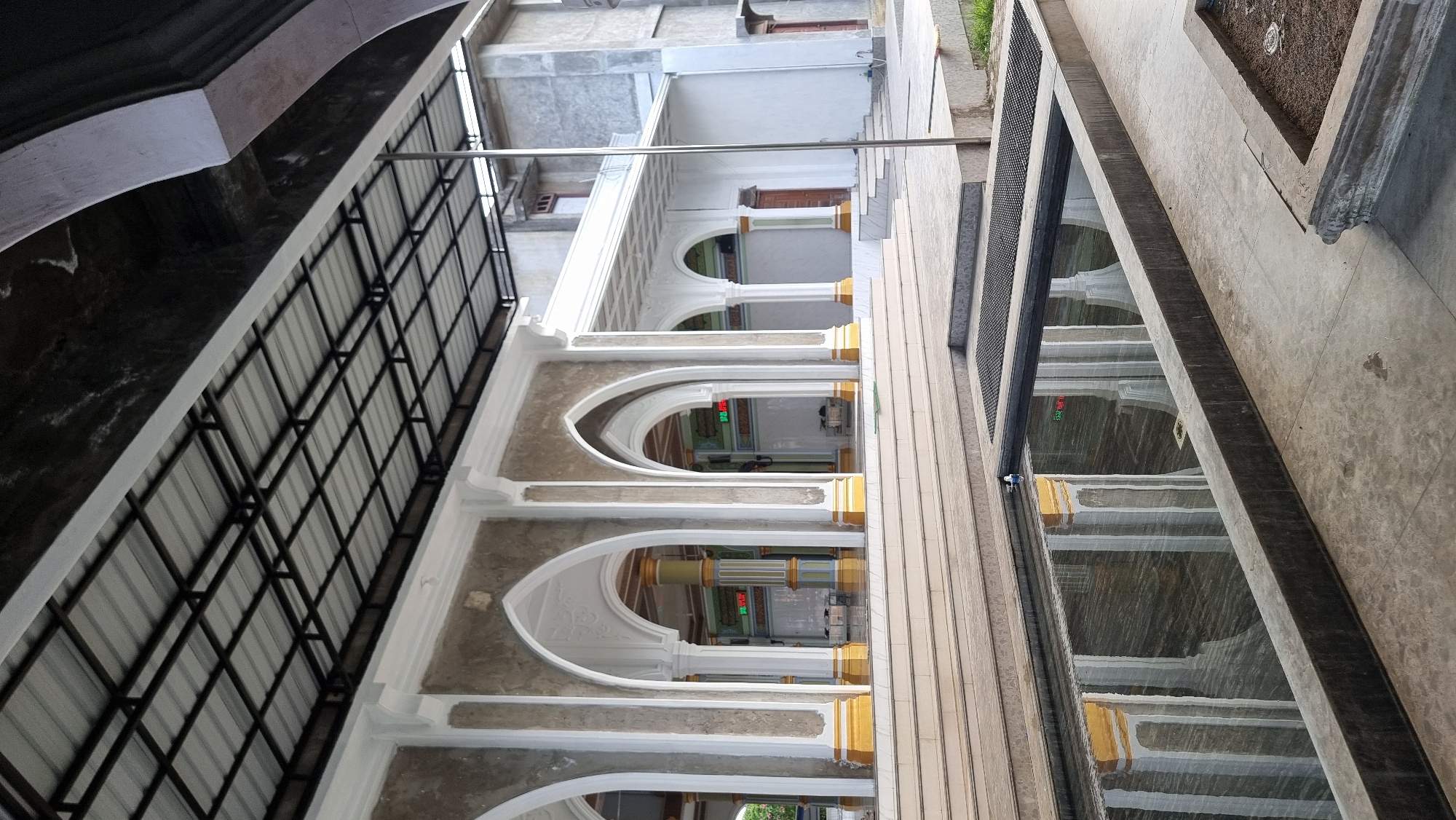 An exterior shot of a mosque in Meunasahlhok, Indonesia, shows a covered walkway with white arched columns, each with a gold accent at the base. The walkway is bordered by a reflecting pool in the foreground. The mosque's facade is light gray with white decorative elements. The roof is a light-colored metal with a black metal frame. A simple metal pole stands to the right of the walkway. The ground is paved with concrete tiles. The sky is not visible.
