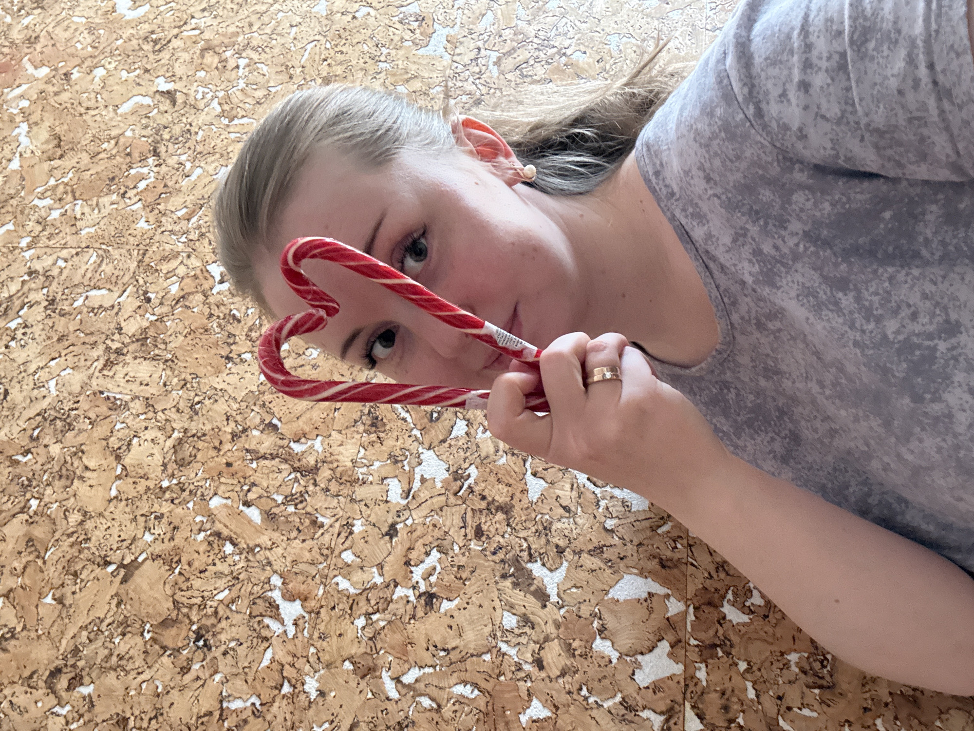 A young adult female is depicted in a close-up, rotated portrait shot, looking directly at the viewer. She has light-colored hair, which appears blonde or light brown, and fair skin. Her expression is neutral. She wears a grey t-shirt with a subtle mottled or speckled pattern and has a ring on her right hand's ring finger. A small, light-colored stud earring is visible in her right ear.
She holds a red and white striped candy cane, which is bent into a heart shape, positioning it in front of her face. Her eyes are visible through the heart shape, and her gaze is directed towards the camera. A small white label with illegible black text is present on the candy cane near where her hand holds it.
The background consists of a textured surface, possibly cork board or a similar material, characterized by irregular pieces of light brown material interspersed with white gaps. The overall impression is playful and direct.