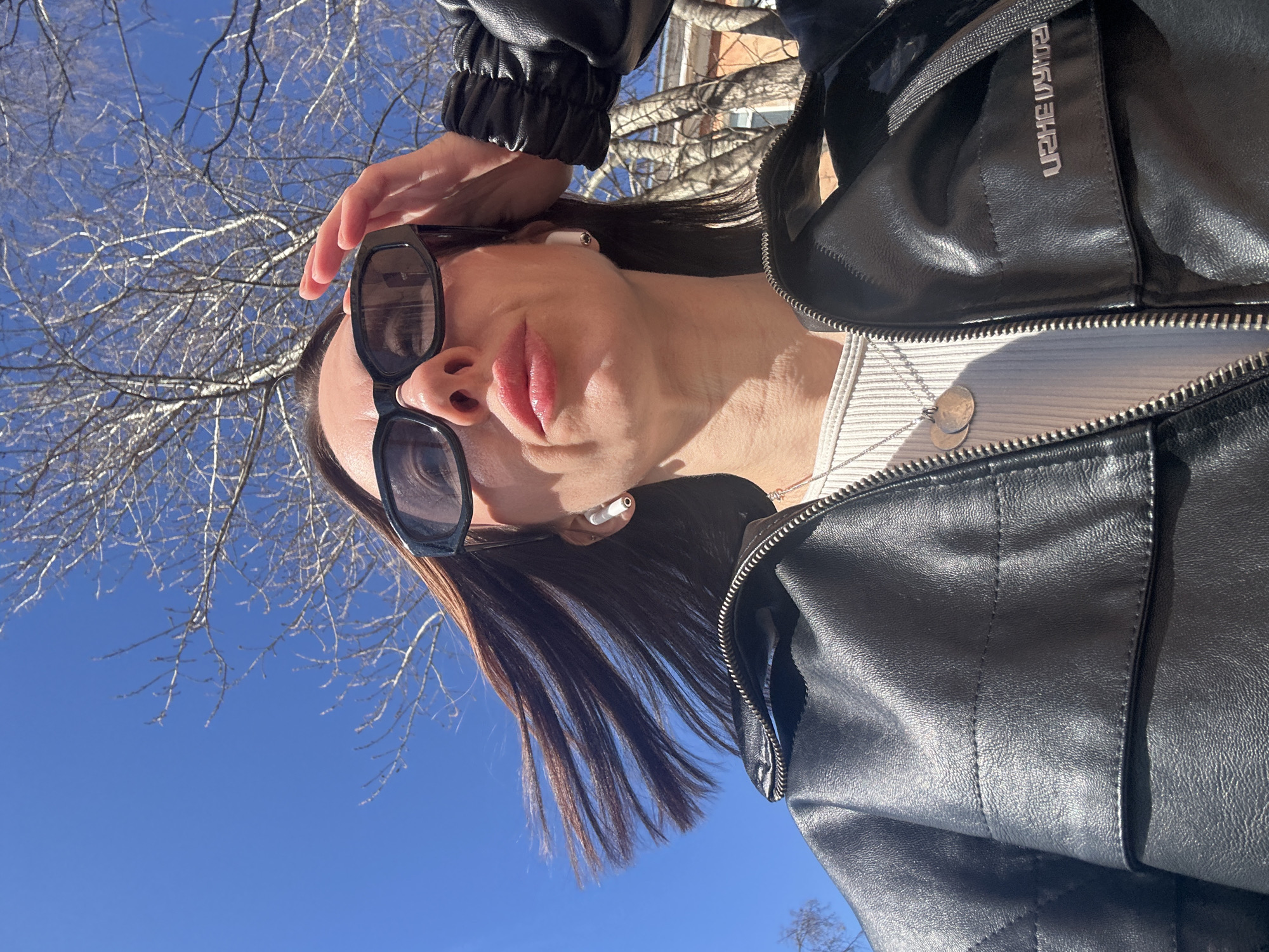 A woman wearing a black leather jacket and sunglasses poses for a photo with the sky and tree branches in the background. She has brown hair, fair skin, and is wearing a white ribbed top under her jacket. The photo is taken from a low angle, looking up at the woman. She is wearing wireless earbuds. The location cannot be determined to be Votkinsk, Russia from the image alone.