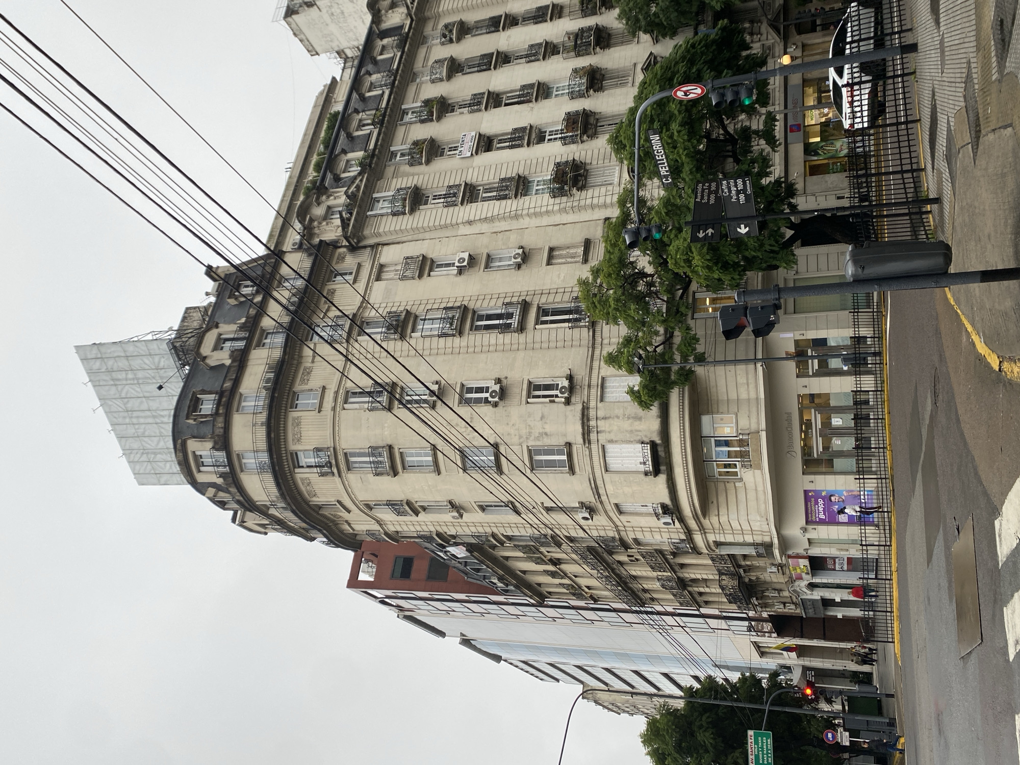 The image depicts a street scene in Buenos Aires, Argentina, showcasing a tall, ornate building with multiple windows and balconies. The building occupies a corner location, curving along the street. The facade is a light beige color, contrasting with the darker metalwork of the balconies. Air conditioning units are visible in some windows. Utility wires crisscross the sky in the foreground.
Street signs indicate "C. Pellegrini" and "Avenida Santa Fe," providing location context. A traffic light stands at an intersection, showing a green light. A Banco Ciudad is visible on the ground floor of the building. Pedestrians and possible parked vehicles are vaguely discernible in the distance on the street. A billboard partially obscures an adjacent building. The overall atmosphere appears overcast.
