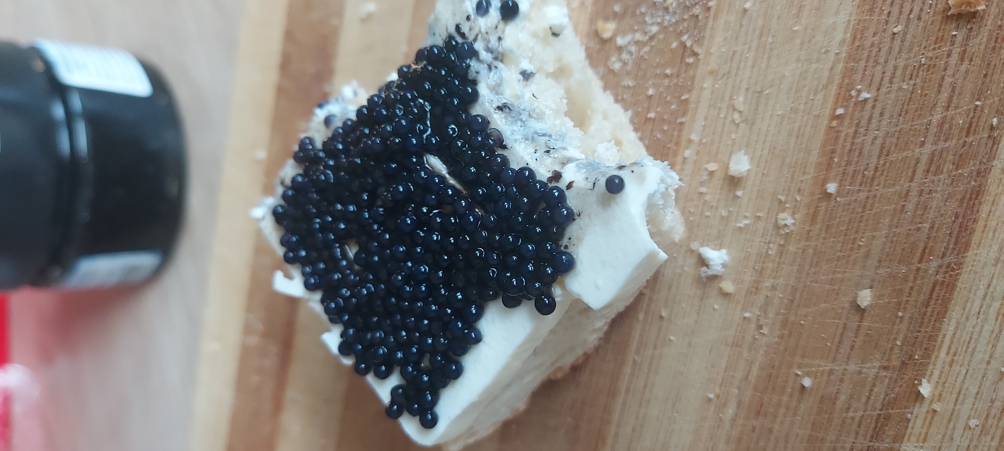 A close-up, high-angle shot shows a piece of bread topped with a thick layer of white spread and a generous amount of black caviar. The bread appears to be a slice of baguette or similar, with its crust visible on the left side and a bite taken out of the bottom right corner, revealing a crumbly interior. The white spread is smooth and opaque, forming a base for the caviar. The caviar consists of numerous small, glistening black spheres densely packed together, with some scattered slightly around the edges. Crumbs of bread are visible on the wooden surface beneath. In the background, a dark jar, likely containing more caviar, is partially visible, along with a blurred red object. The scene is set on a light-colored wooden cutting board. There is no indication of the specific location in Castelldefels, Spain.