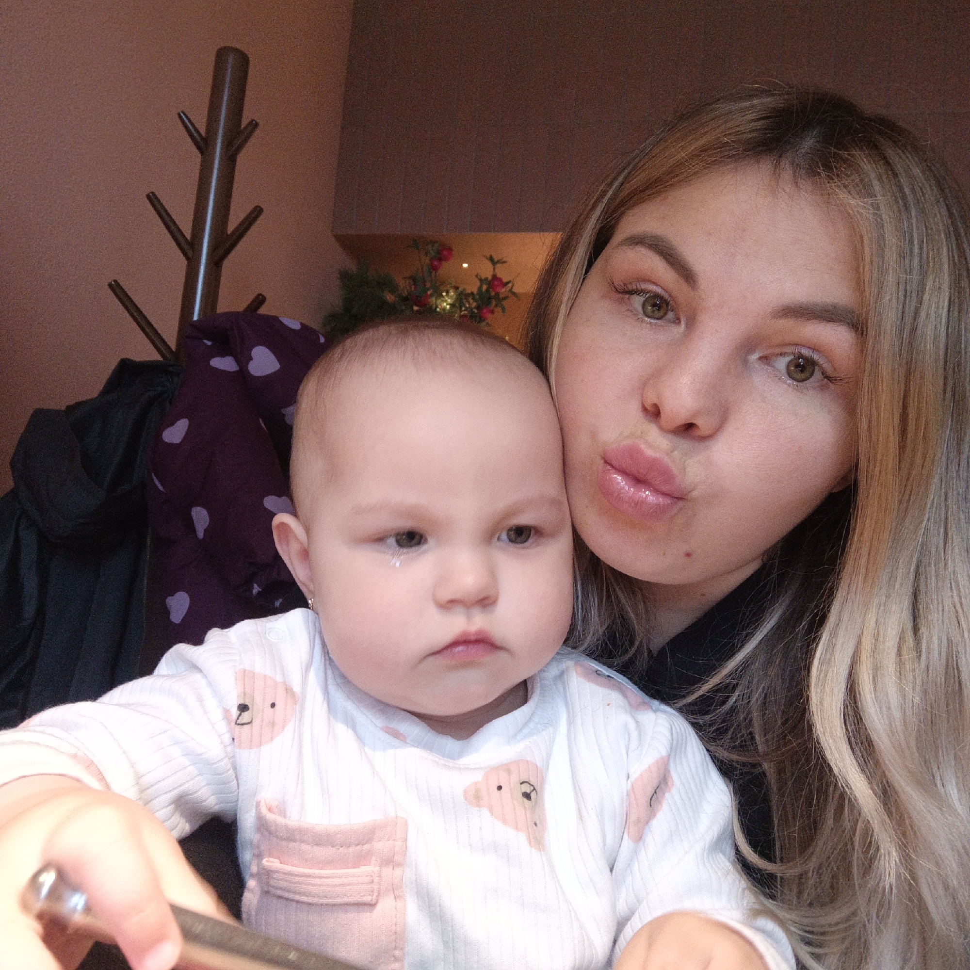 A selfie photograph shows a blonde woman with a baby. The woman is on the right, making a kissy face with glossy lips. The baby, wearing a white onesie with pink bear designs and a pocket, appears to be crying and has a tear rolling down its cheek. The setting appears to be indoors, with a coat rack, a jacket with a heart pattern, and some greenery with red berries visible in the background. The city and location are unknown.