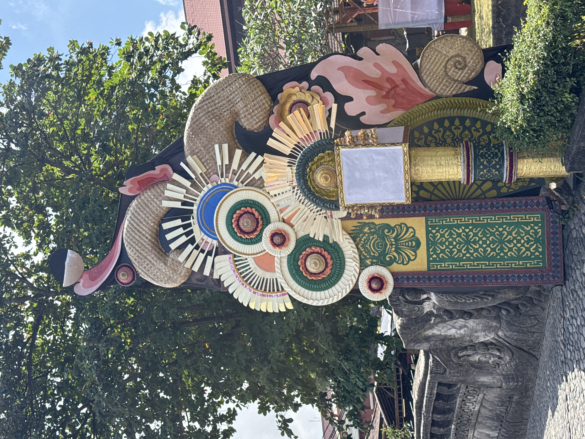 The image displays a vibrant and intricate cultural decoration, likely part of a festival or ceremony in Denpasar, Indonesia. The main subject is a large, elaborate adornment featuring a variety of textures and colors. Woven straw elements, resembling coiled snakes or stylized horns, are prominently featured, along with circular fan-like designs made from layered materials like palm leaves and fabric. These circular elements have detailed patterns in shades of green, blue, white, orange, and pink.
A gold-framed rectangular object, possibly a small screen or decorative panel, is situated at the center of the arrangement. The entire decoration is affixed to a dark background, accented with stylized floral or flame-like shapes in pink and red. Below the central decoration, a long, ornate band displays repeating patterns in green and gold, bordered by red and blue trim, and further embellished with a mosaic of small, colorful woven circles.
The background shows lush green foliage from trees, suggesting an outdoor setting. The sky is visible with some white clouds, indicating it's daytime, likely bright. Parts of a stone structure, possibly a temple or an ancient building with carved details resembling a dragon or mythical creature, are visible at the bottom and right of the frame, providing a strong cultural and locational cue. The overall impression is one of festive celebration and rich cultural heritage.